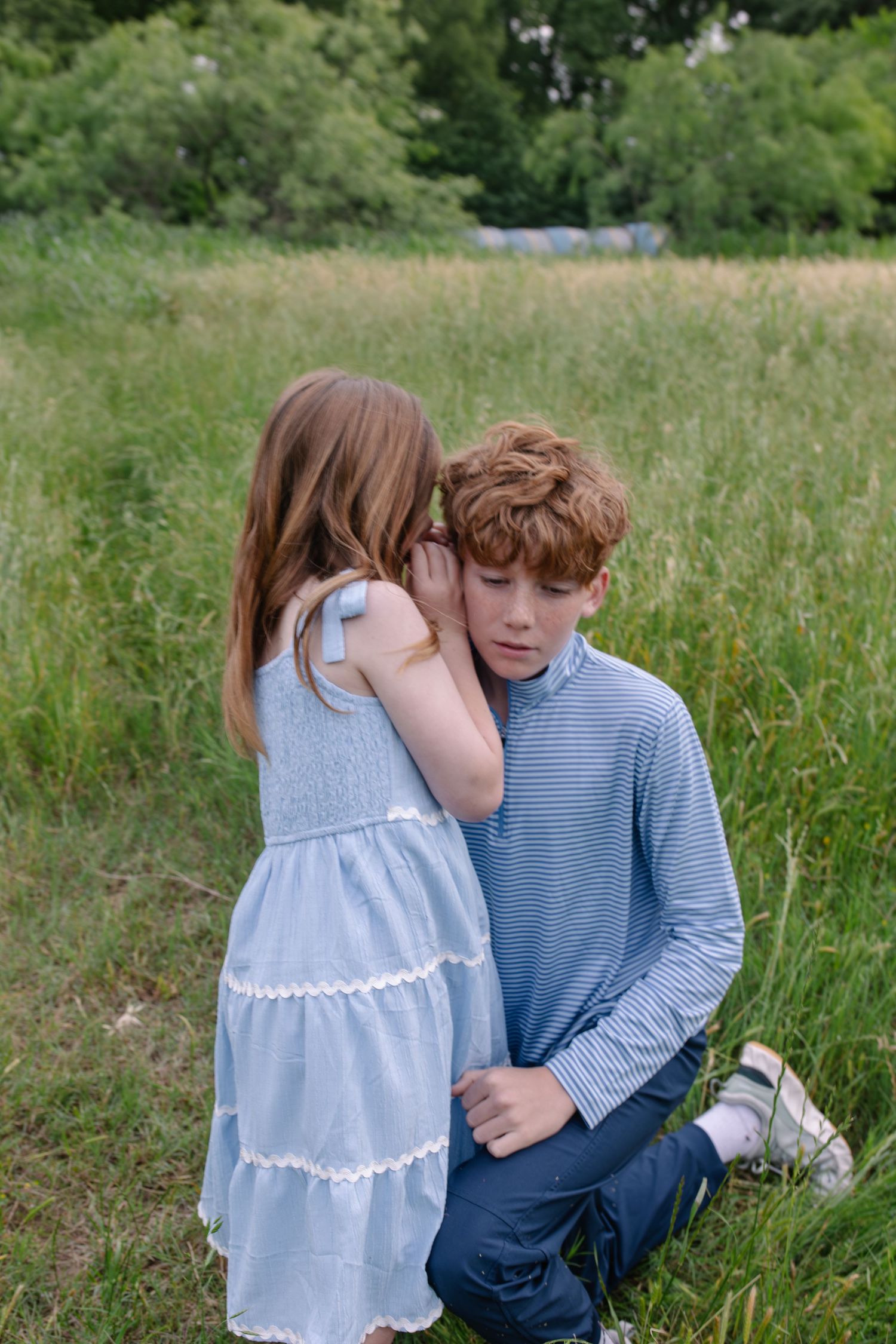 A young girl in a blue dress whispers to a red-haired boy kneeling in a lush green meadow.
