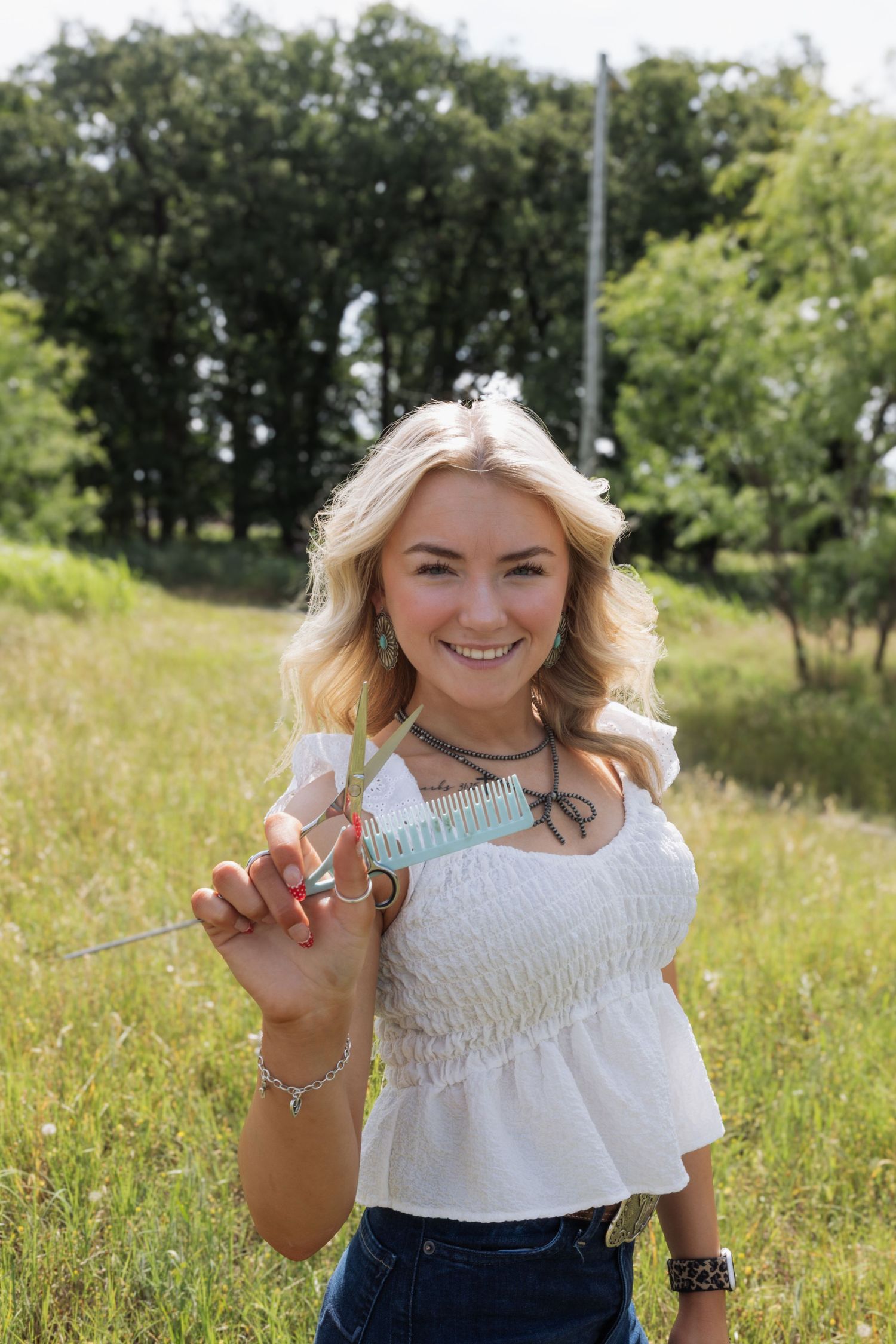 Smiling blonde woman in white top holding a decorative skewer in a green outdoor setting.