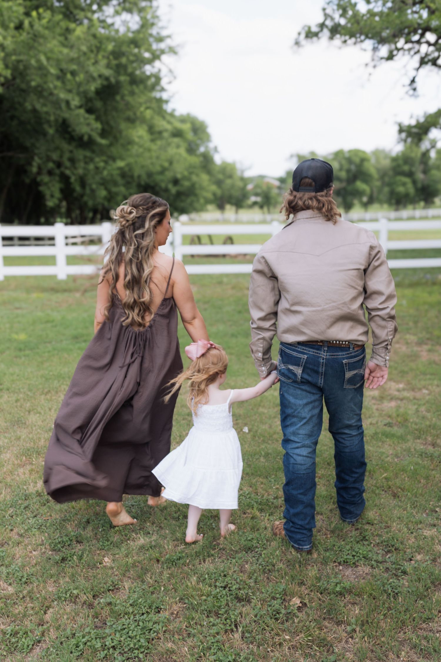 A family of three walks hand-in-hand through a green pasture, with a white fence and trees in the background.