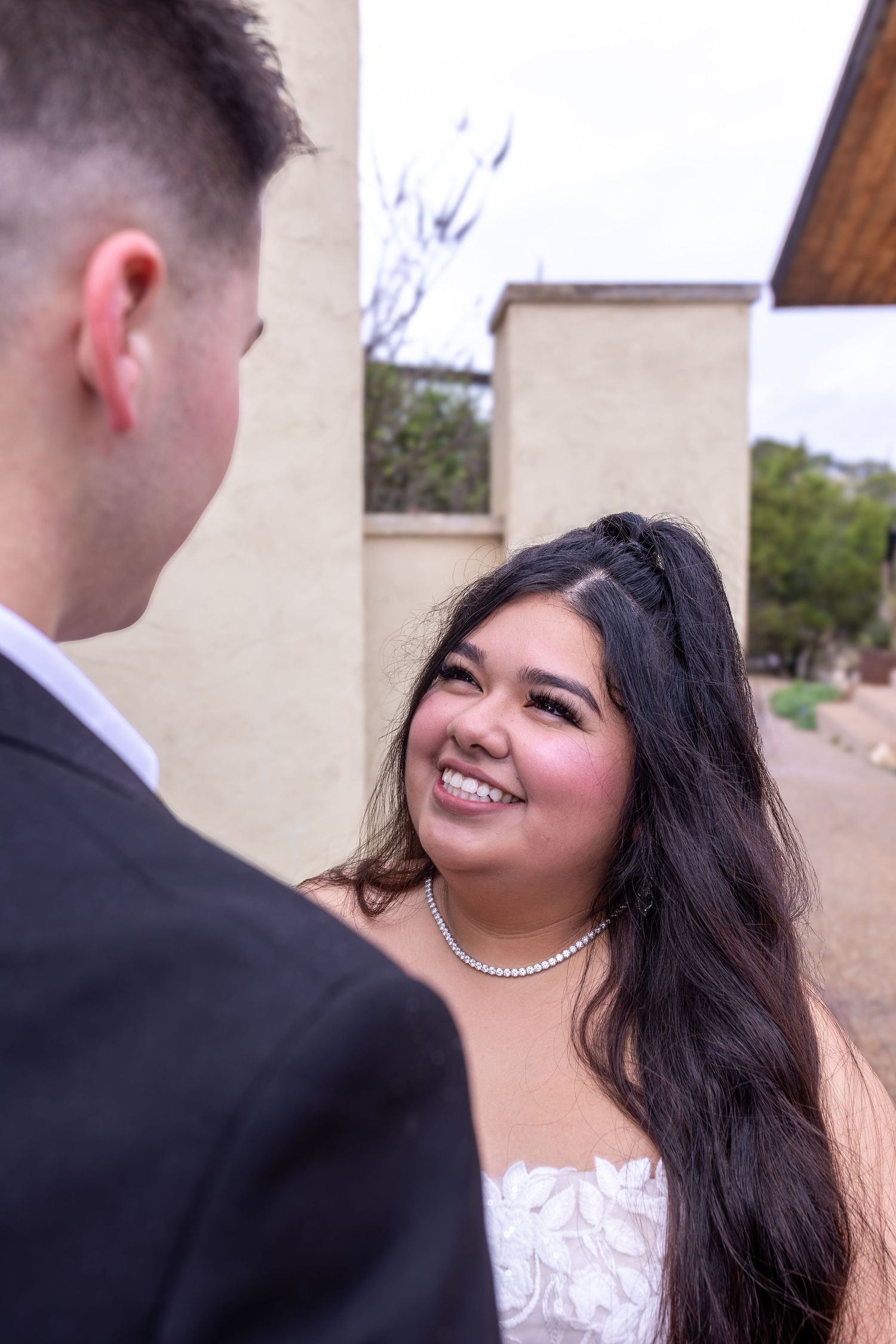 A couple shares a romantic moment during their wedding photoshoot, with the bride wearing a white lace dress and pearl necklace.