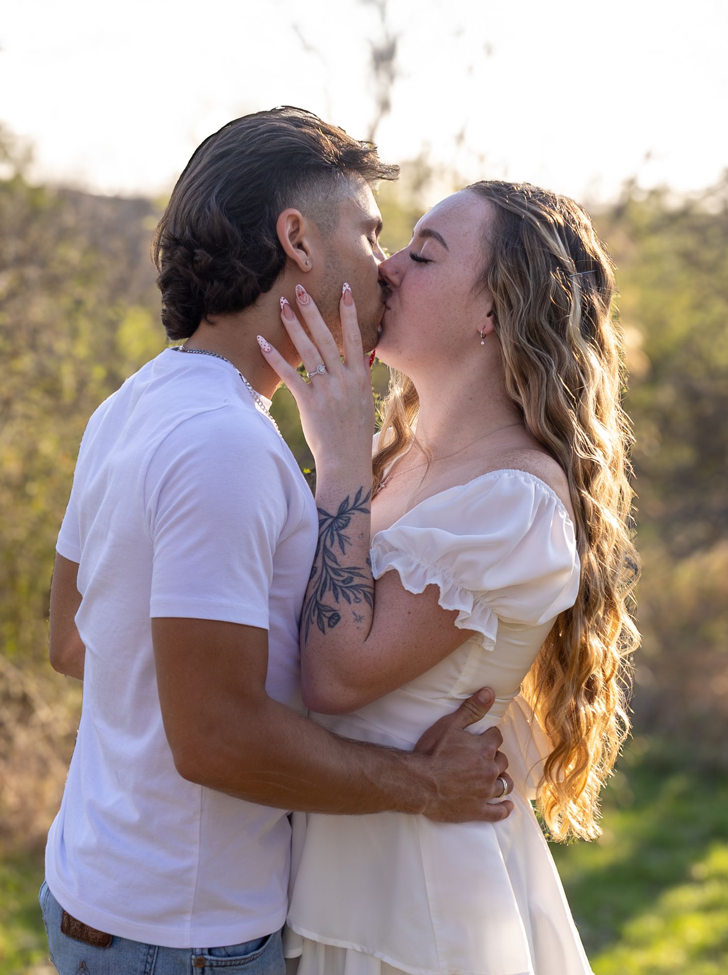A couple in white outfits share romantic kisses in a sunlit outdoor setting with natural greenery in the background.