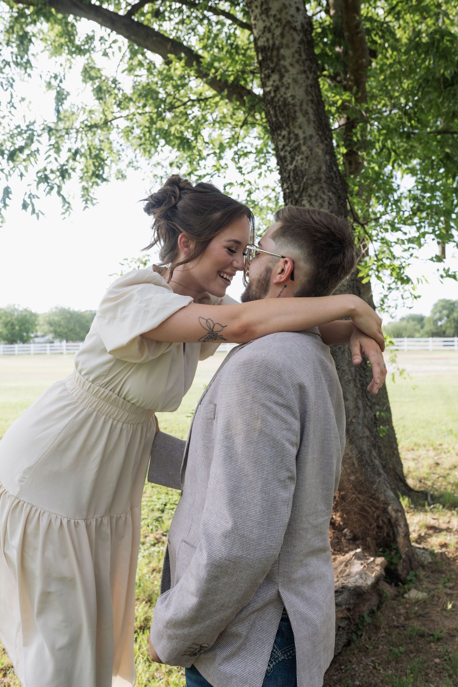 Couple sharing a romantic moment under a tree in a park, woman in white dress embracing man in gray jacket.