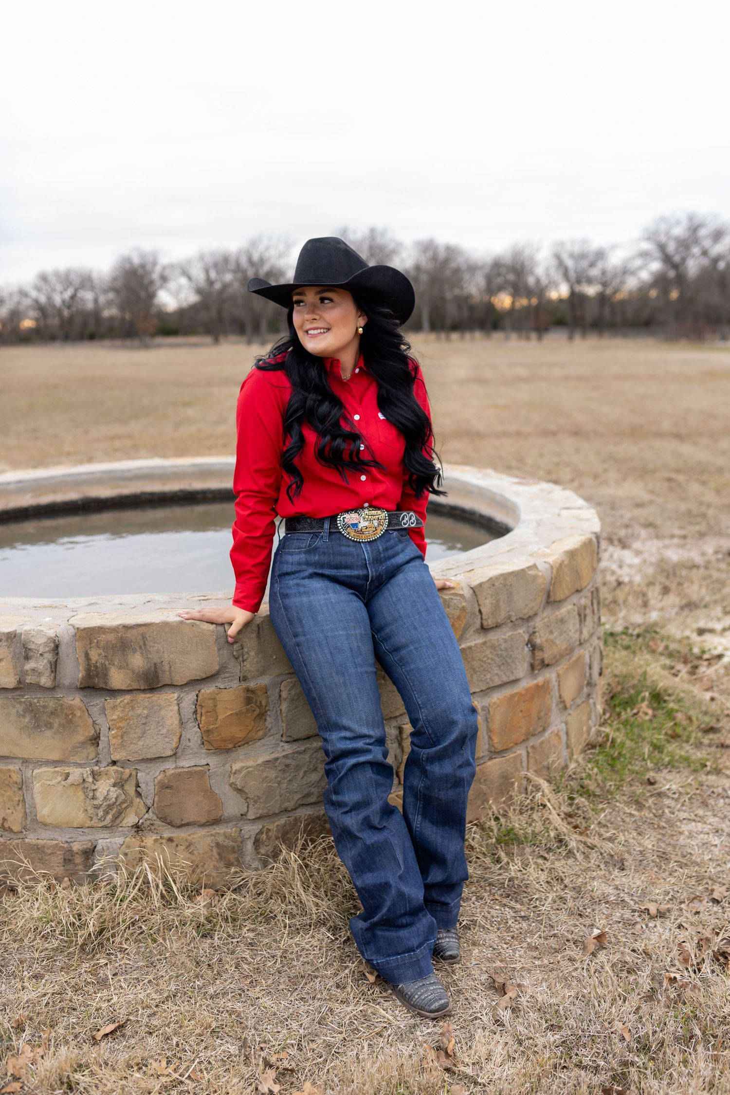 Country western outfit with red top and jeans sitting on stone wall in rural field setting.
