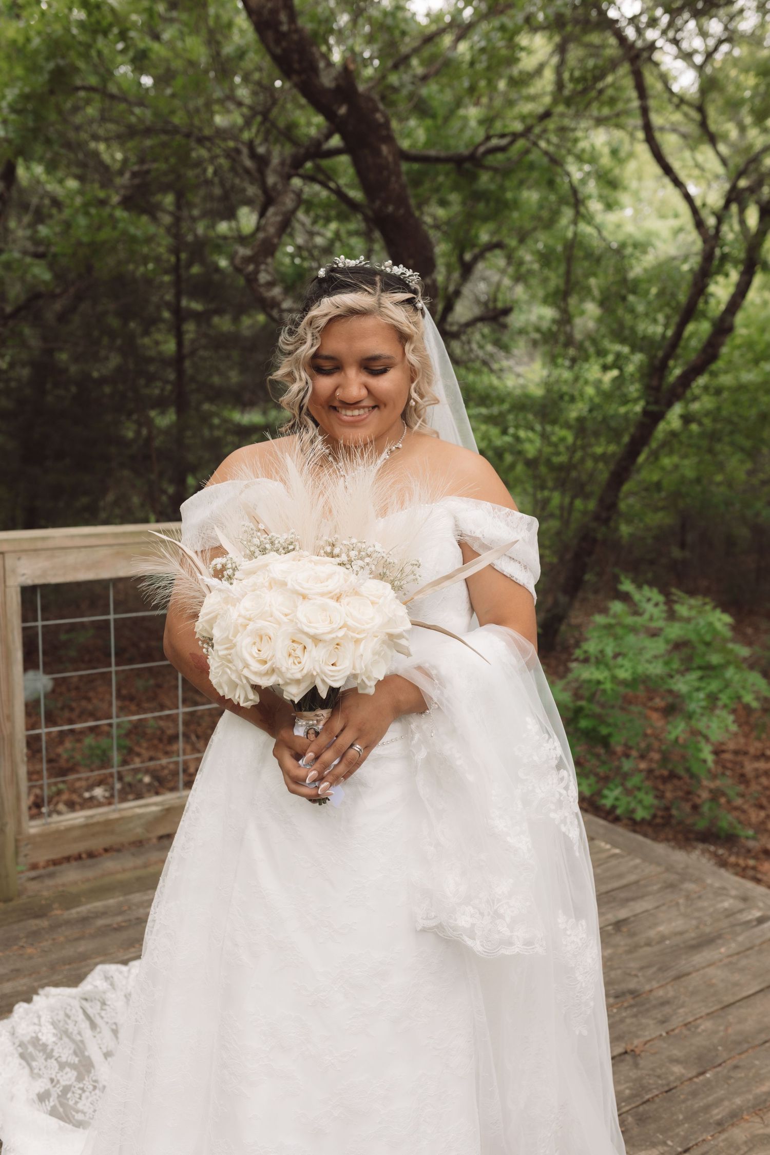 Smiling bride in off-shoulder white lace gown holds white rose bouquet on wooden deck surrounded by lush greenery.