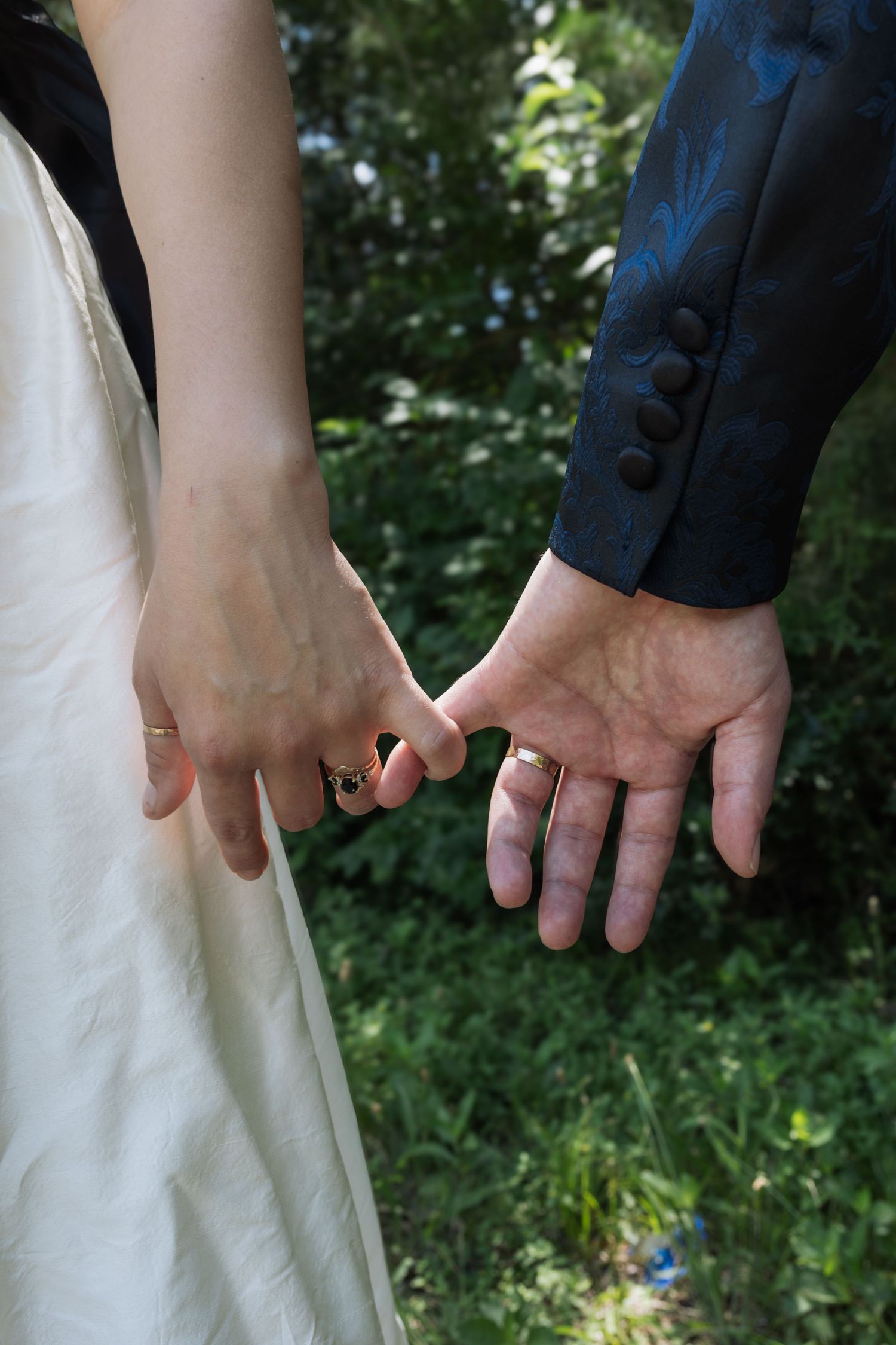 Close-up of newlywed couple holding hands, showing wedding rings, bride in white dress, groom in blue suit, green foliage background.
