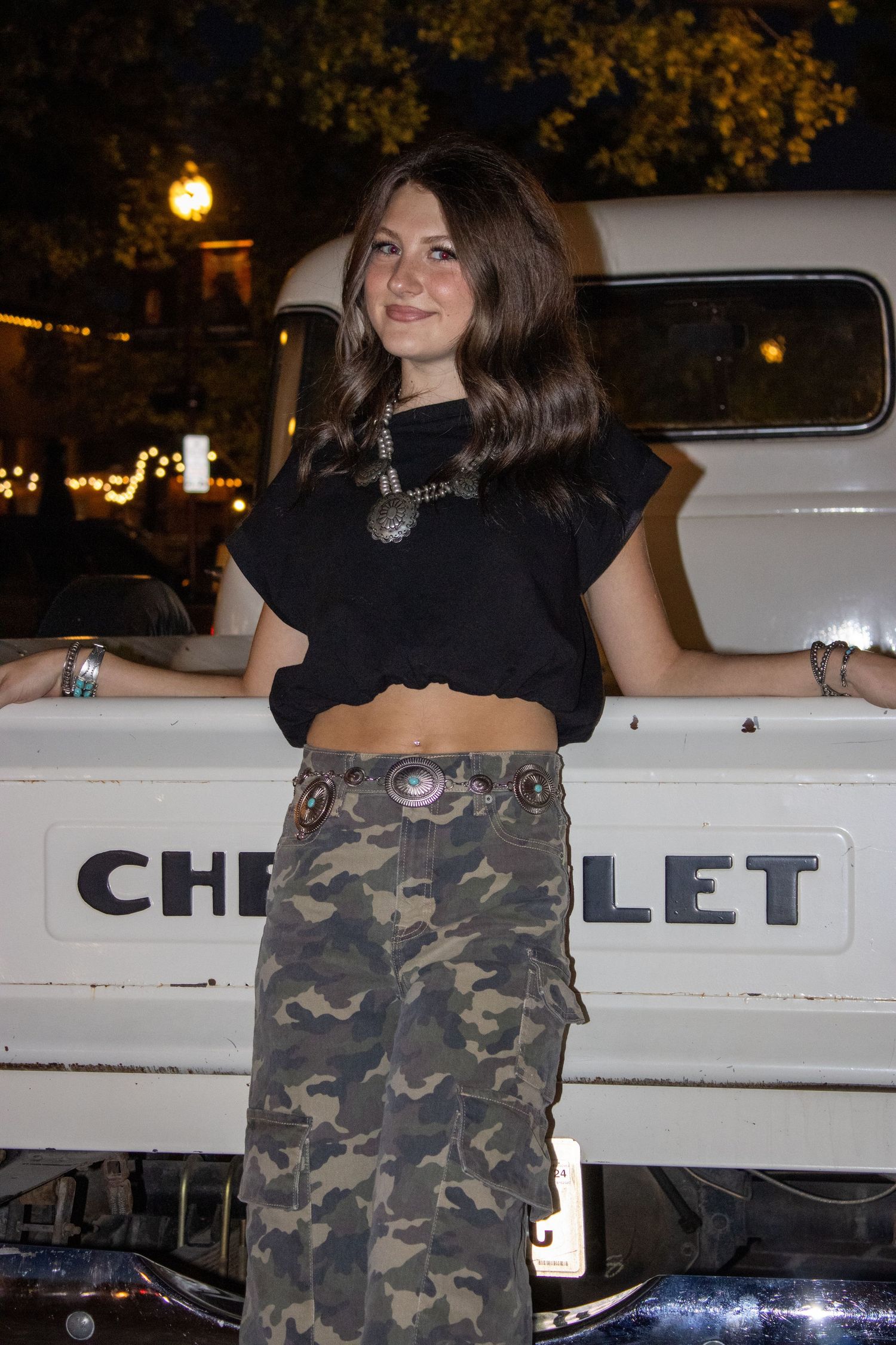 Person in black top and camouflage pants posing near white Chevrolet truck.