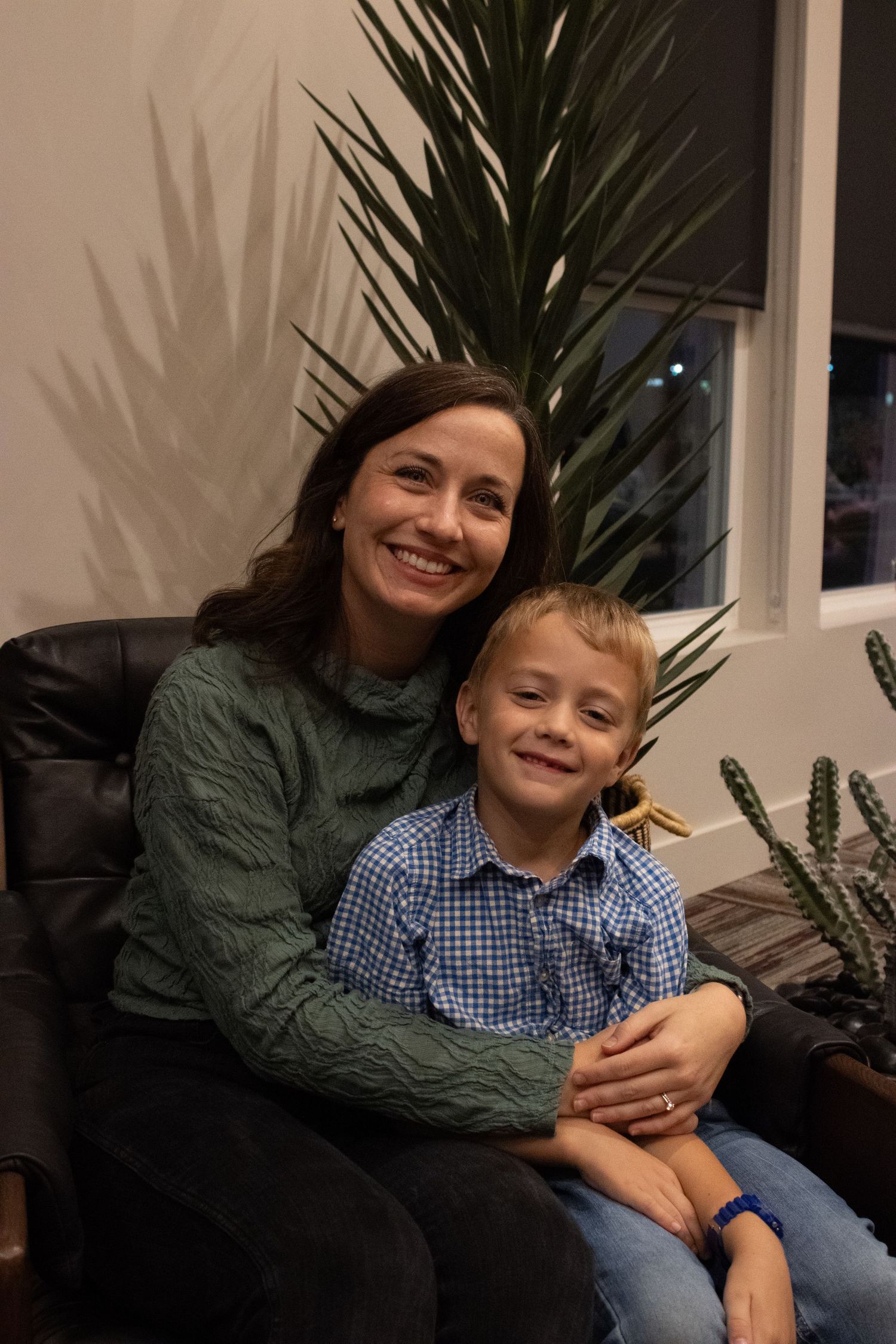 A warm indoor portrait of two people sharing a moment on a leather chair beside a tall palm plant.