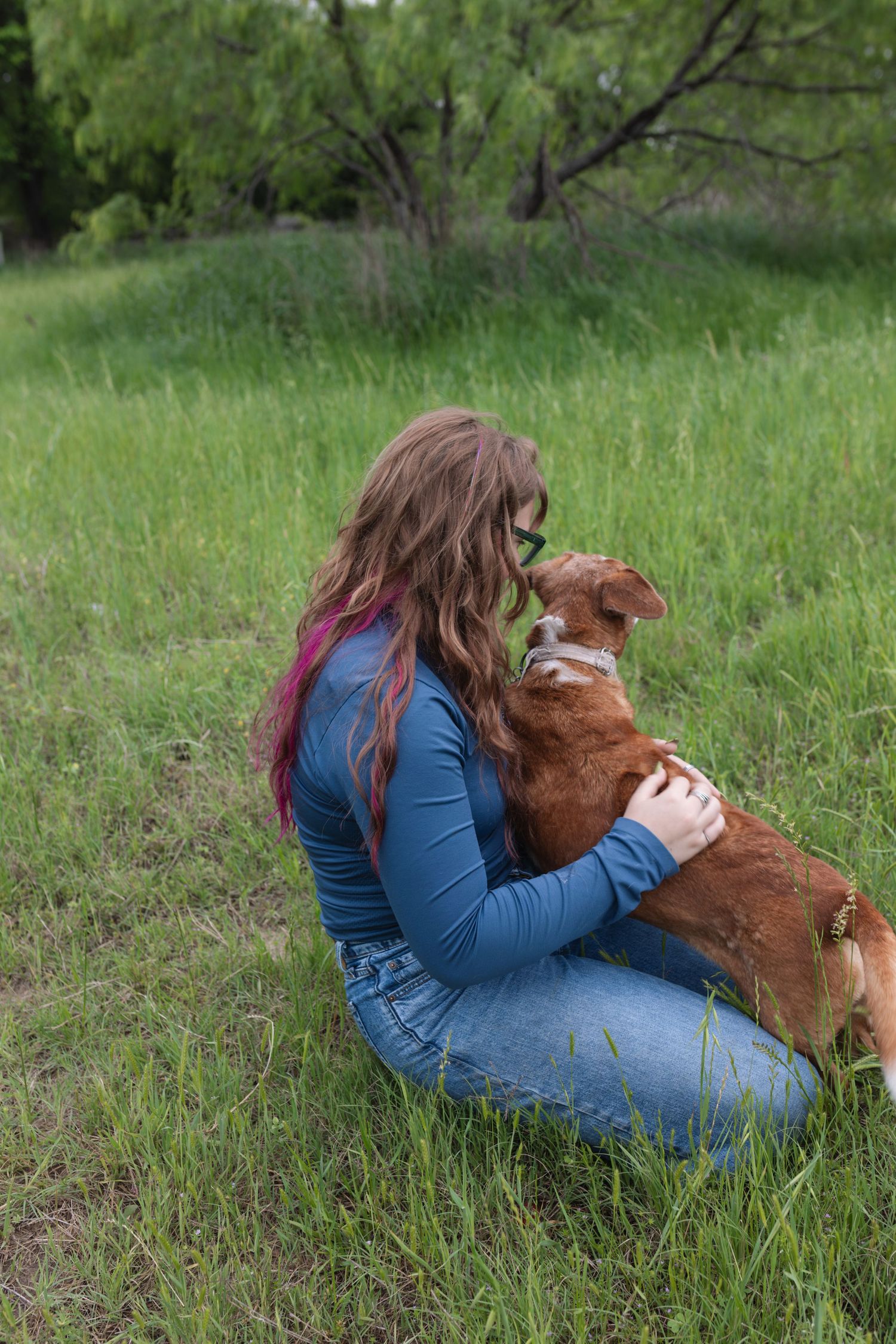 Young woman with pink highlights sits in green grass hugging her brown dog outdoors.