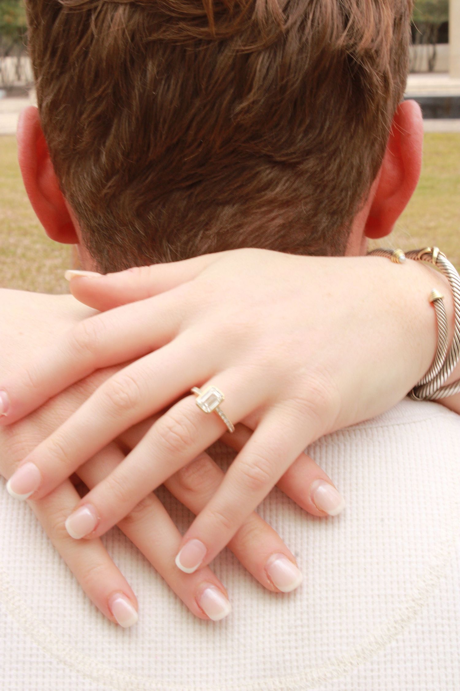 Woman's hand with emerald cut diamond engagement ring and bracelet resting on man's shoulder.
