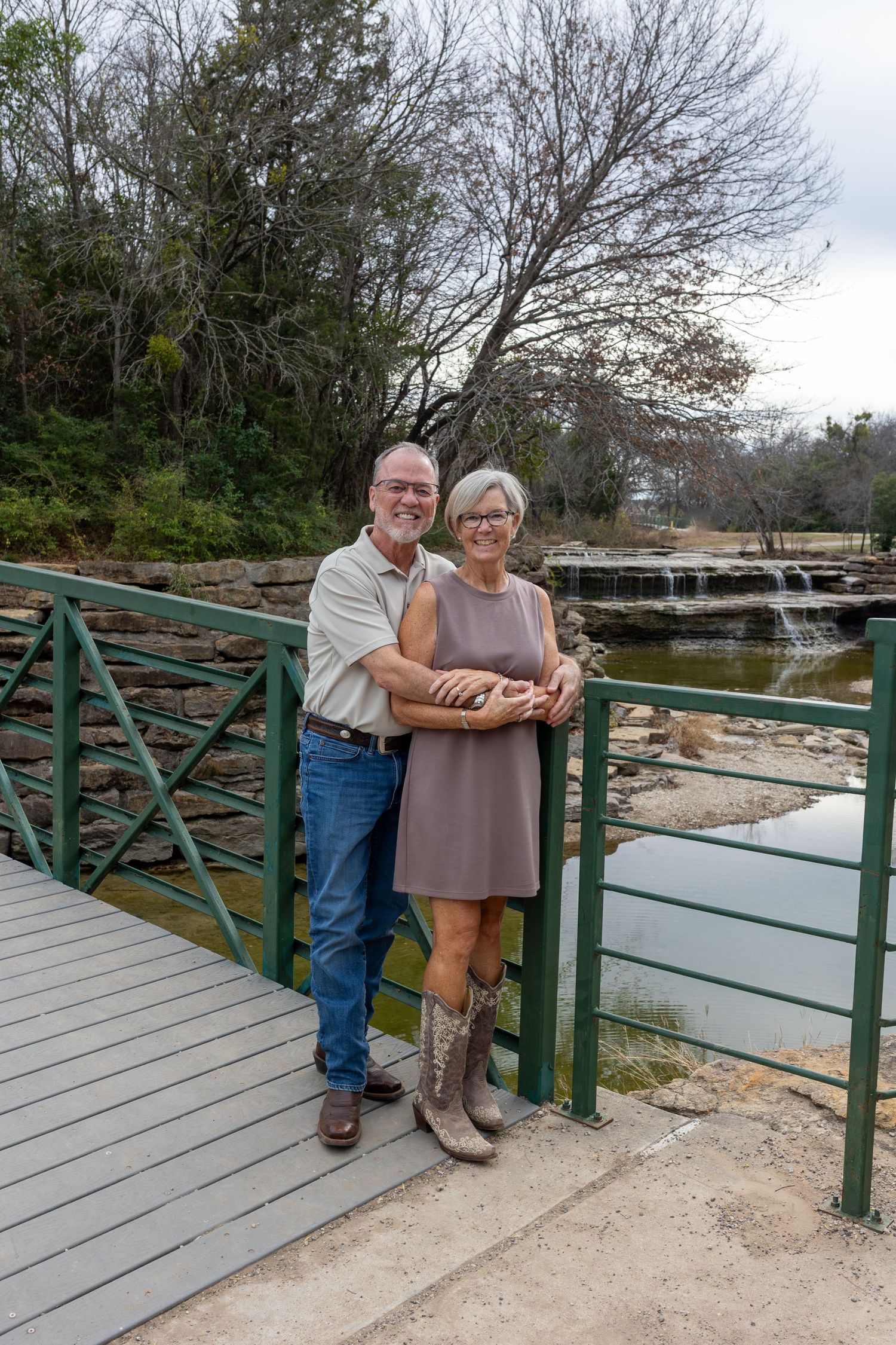 A couple stands together on a wooden bridge with green railings on an overcast day.