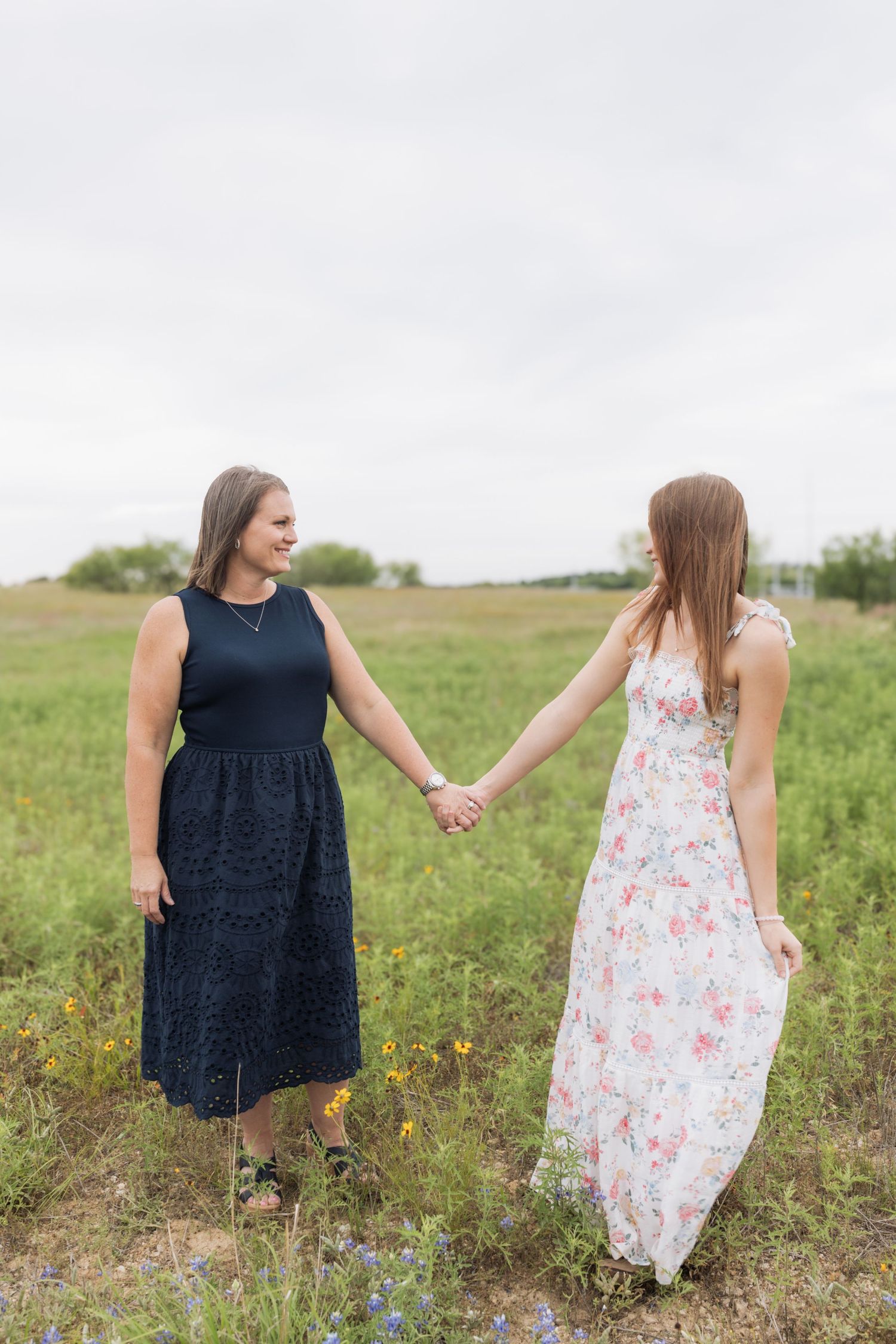 Two women in dresses hold hands smiling in a green field.