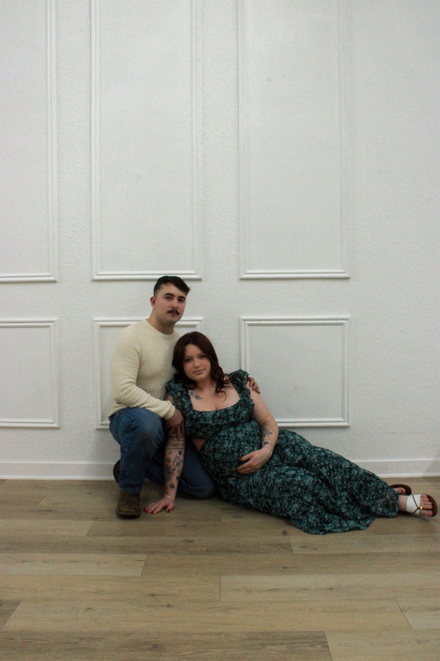 A couple reclines together on a hardwood floor against a white wall with decorative molding.