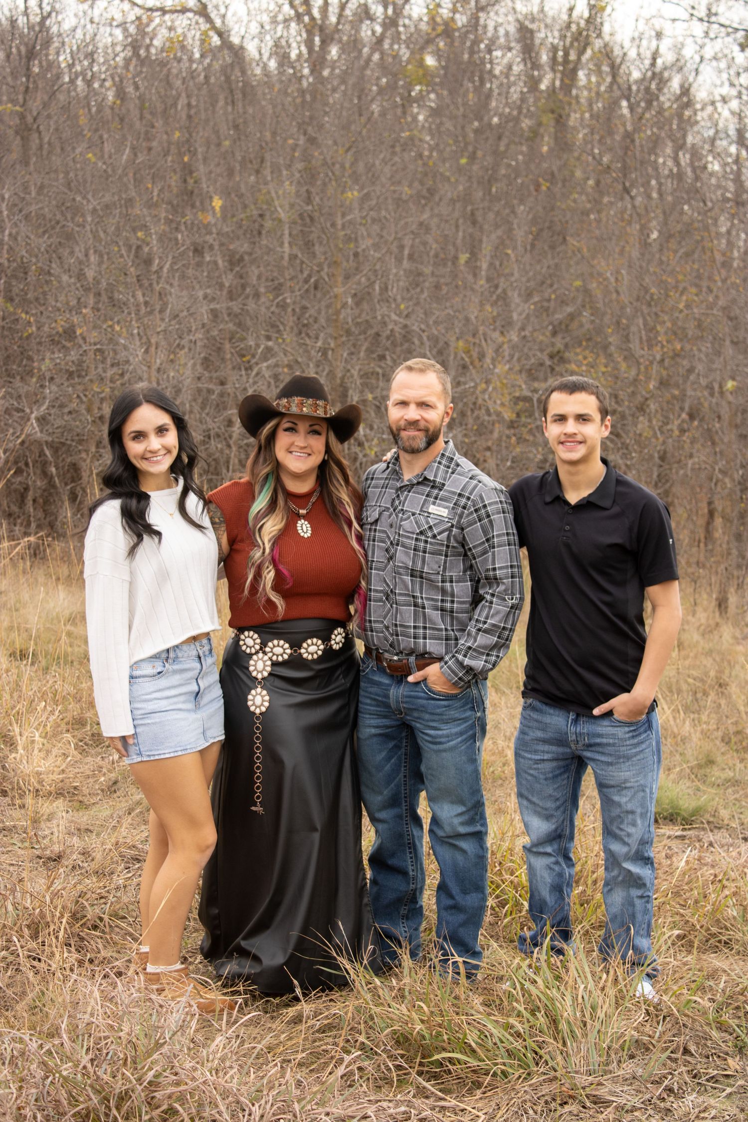 A casual outdoor family photo shoot with people lined up in front of brown winter foliage and trees.