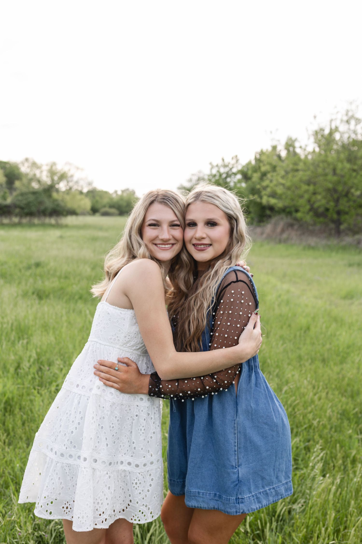 Two smiling blonde young women hugging in a green field, wearing white and denim dresses.