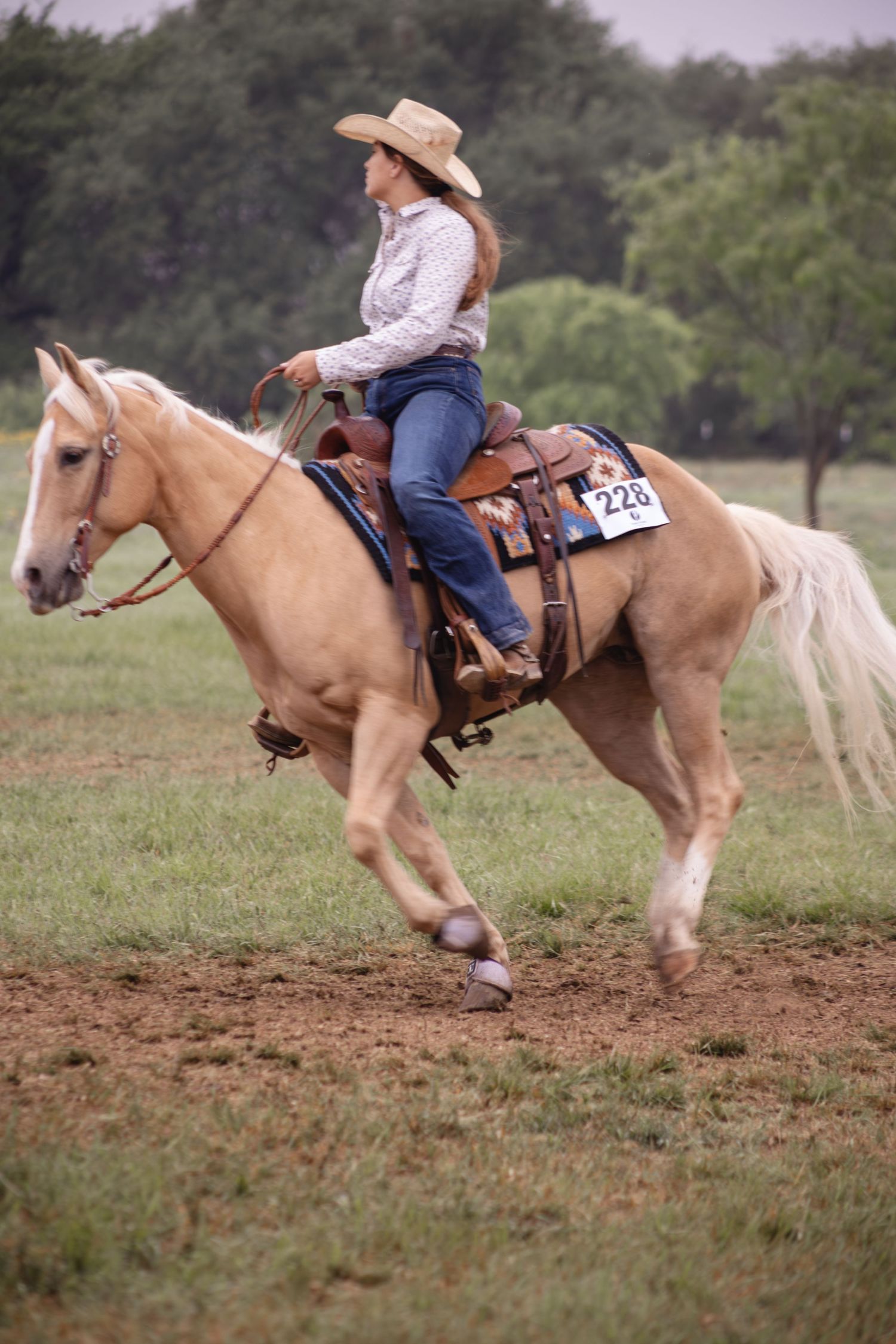 Cowgirl in straw hat rides palomino horse numbered 228 at outdoor equestrian competition on dirt field.