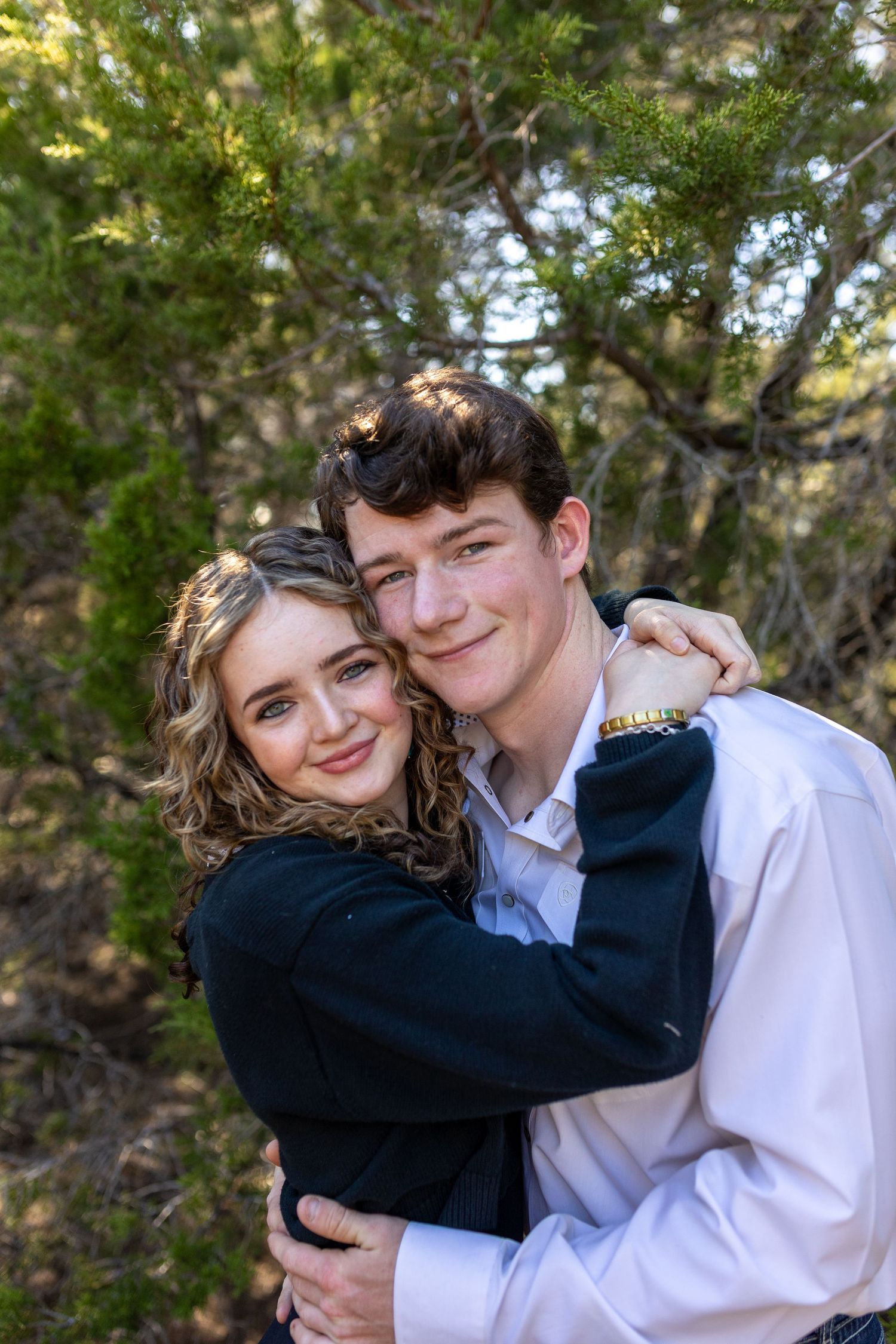 A young couple embracing outdoors, smiling warmly, with green trees in the background, dressed in formal attire.