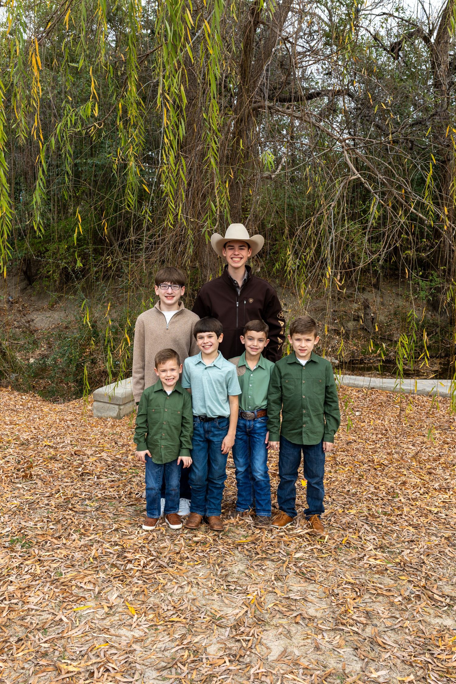 Children lined up outdoors on fallen leaves with trees and foliage in the background.
