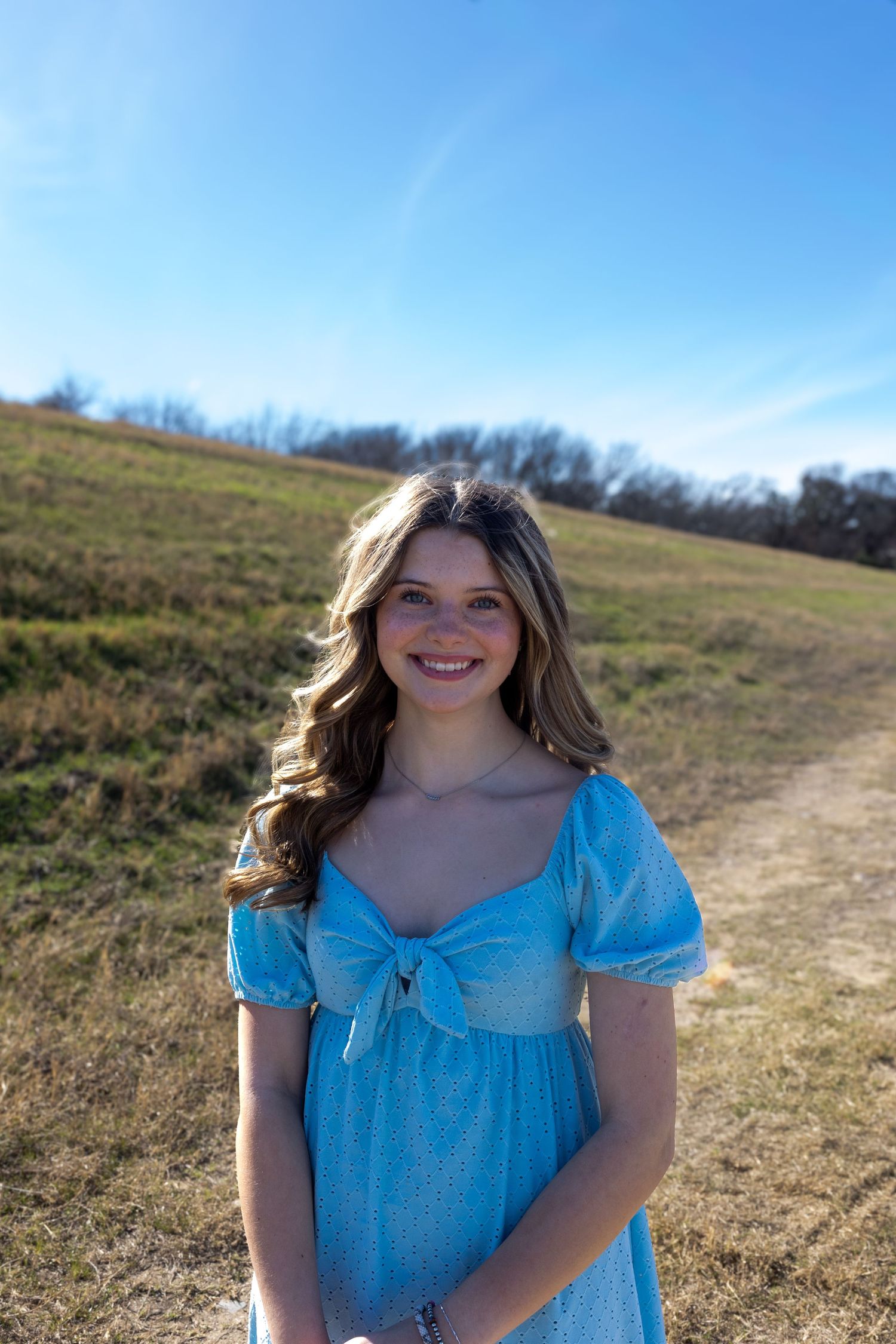 A person in a light blue dress poses on a grassy hillside on a sunny day.