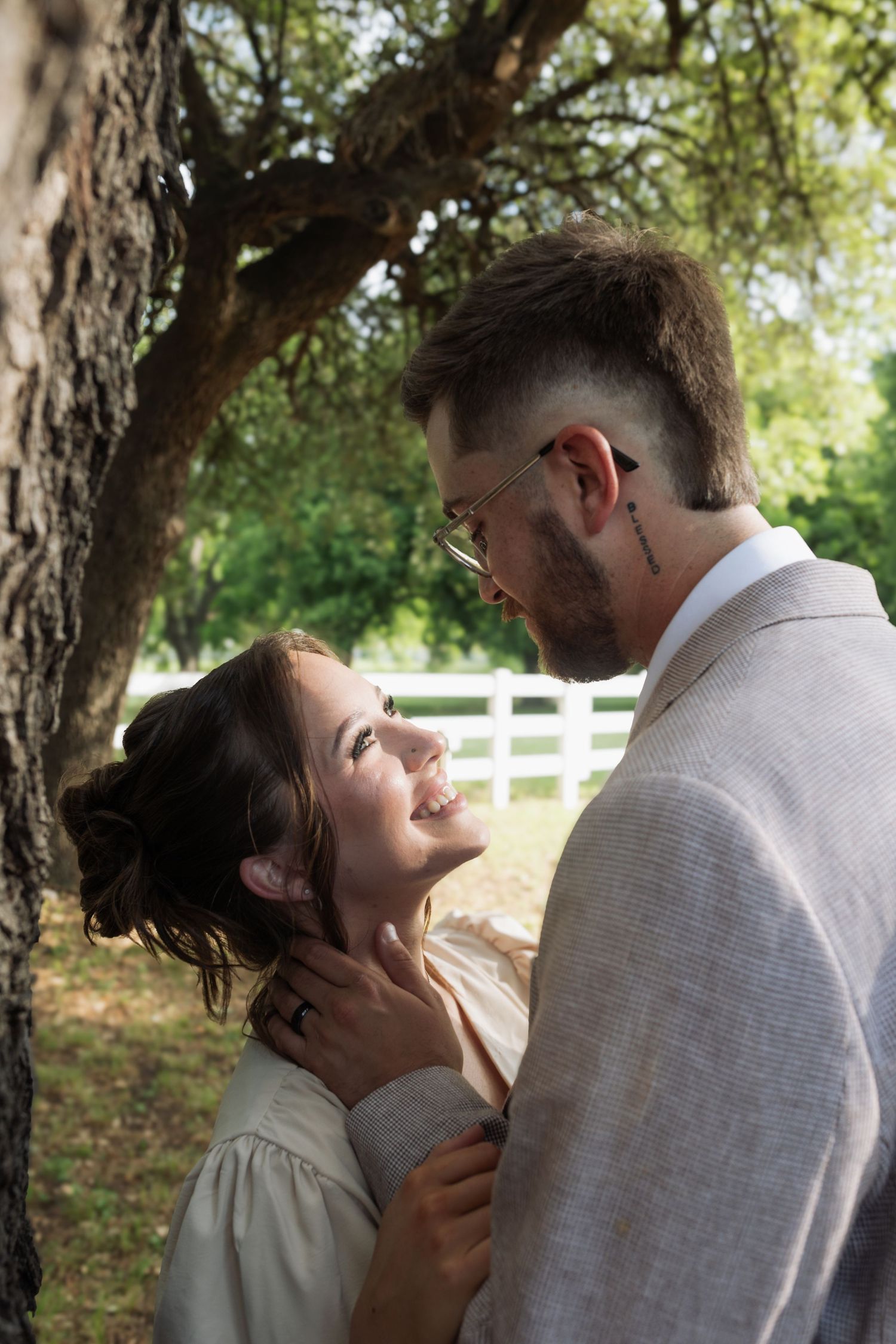 Romantic couple embracing under a tree outdoors, woman smiling up at man wearing glasses near white fence.