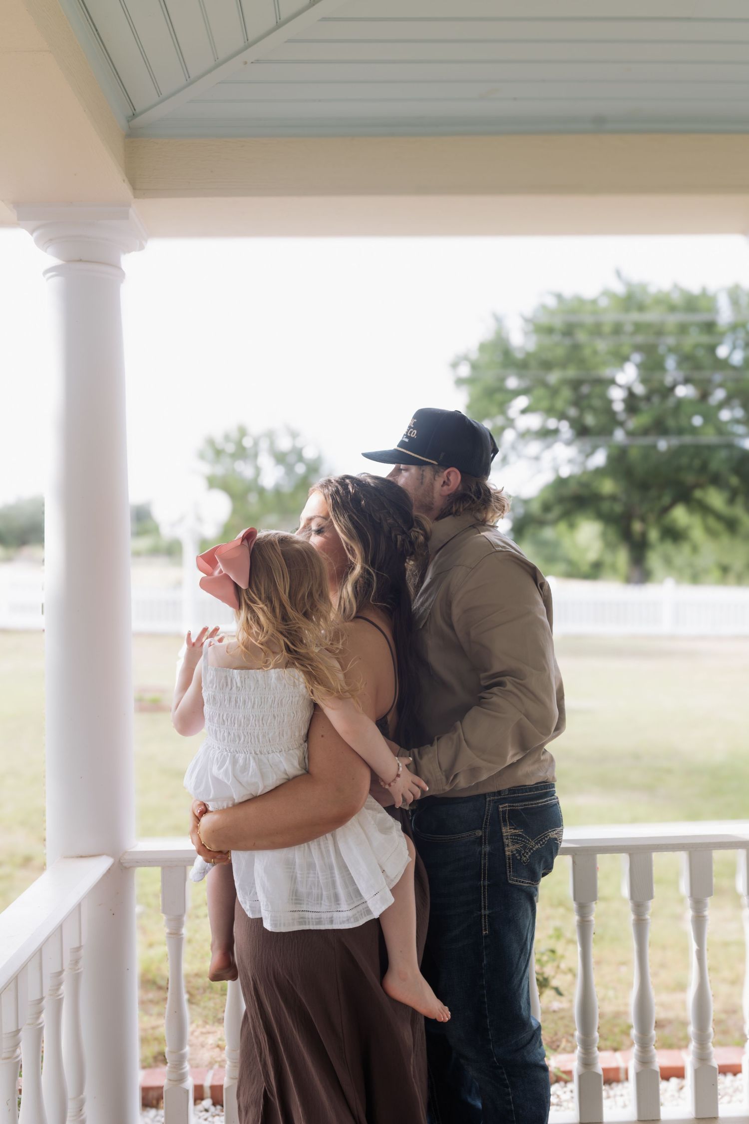 Family sharing a tender moment on a farmhouse porch, mother holding child while father kisses her head, rural setting.
