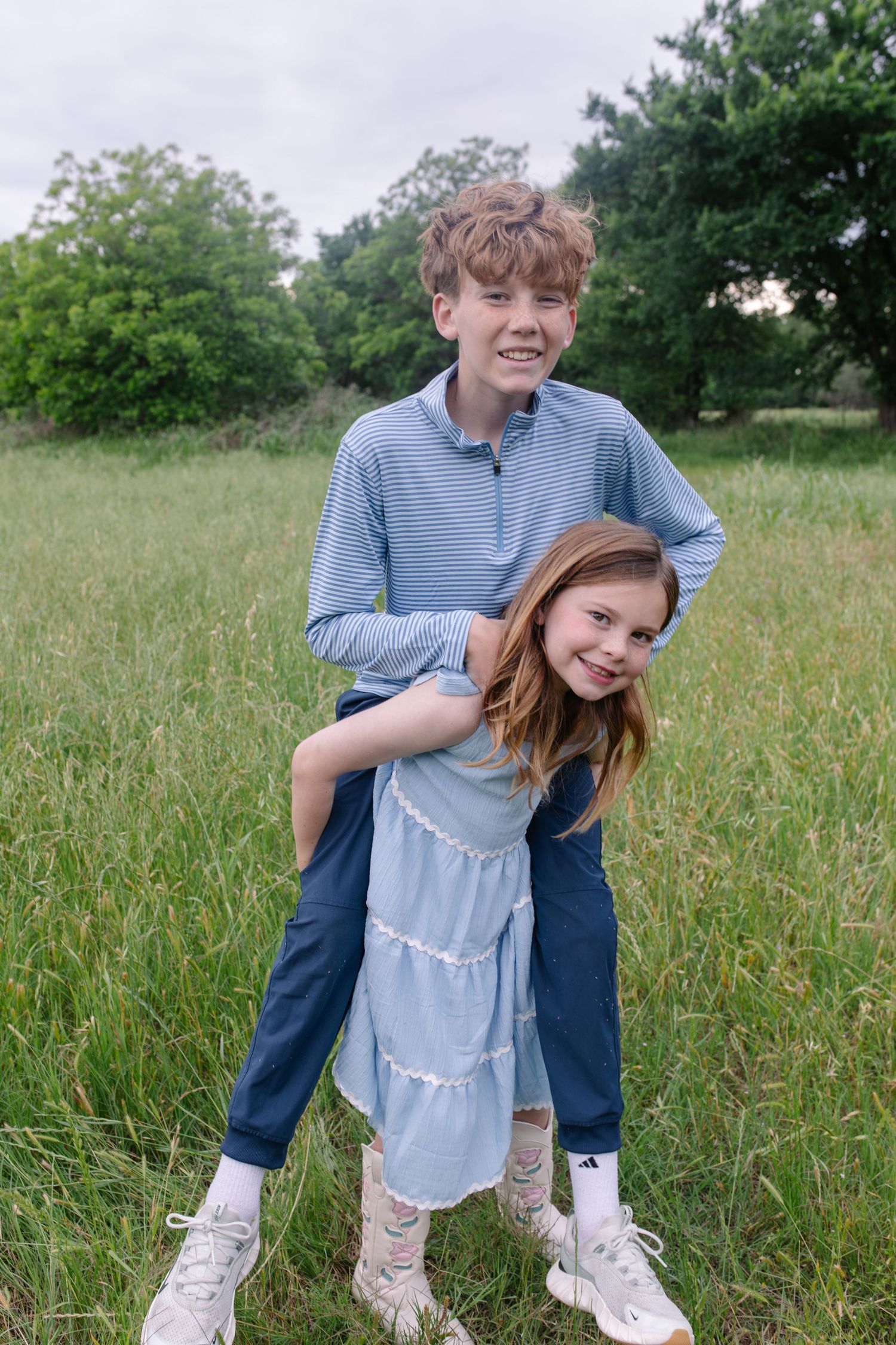 Two smiling children in blue outfits play in a green field, with an older boy hugging a younger girl from behind.