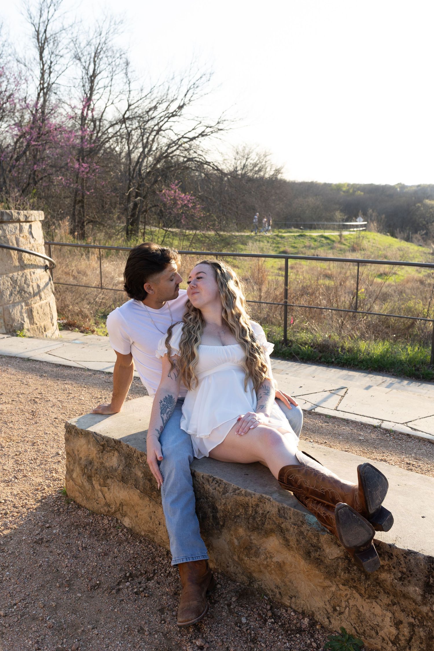 A romantic couple sits on a stone wall during sunset with blooming trees and a rural landscape in the background.