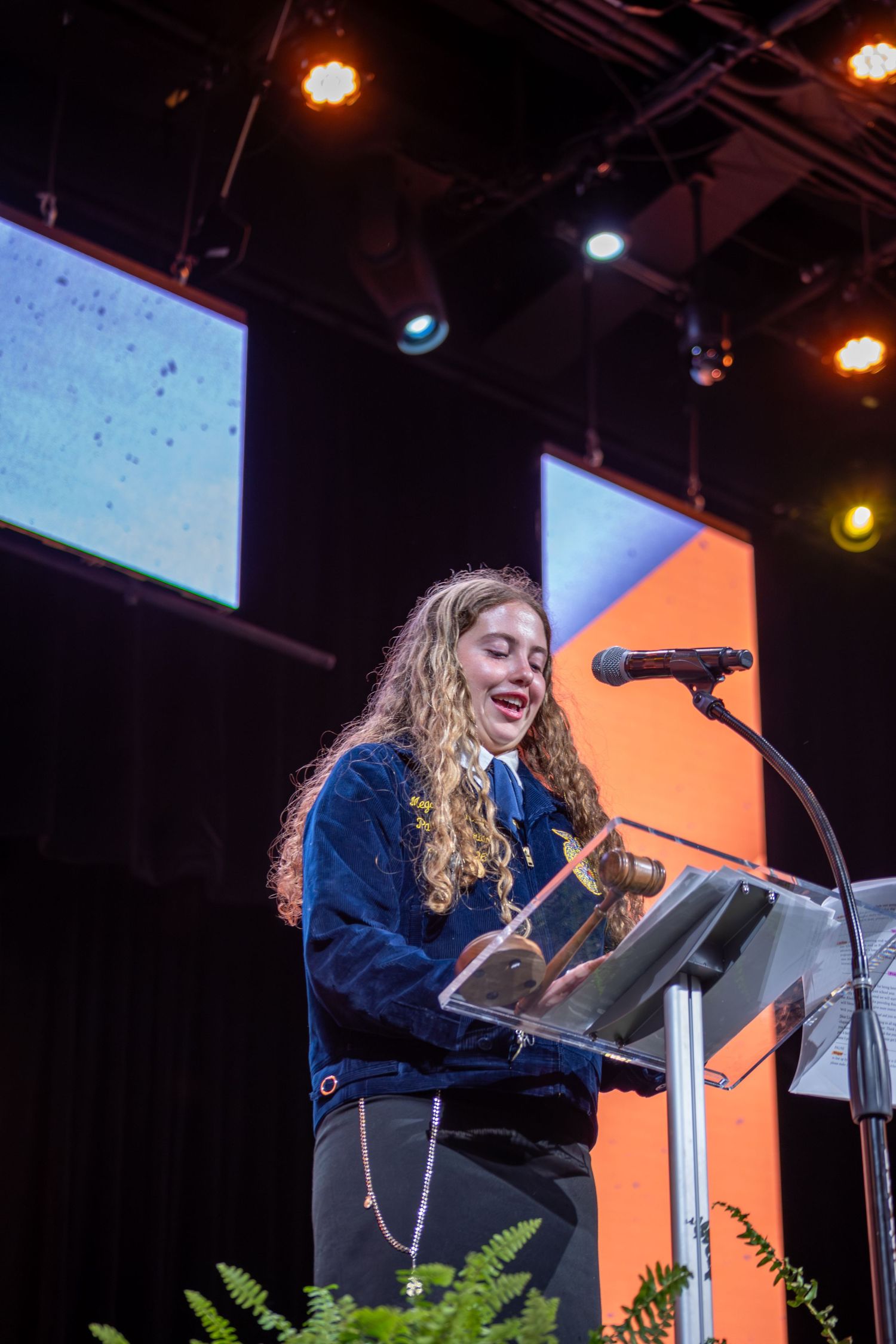 Young girl in FFA jacket speaks at podium with microphone on stage with colorful screens behind her.