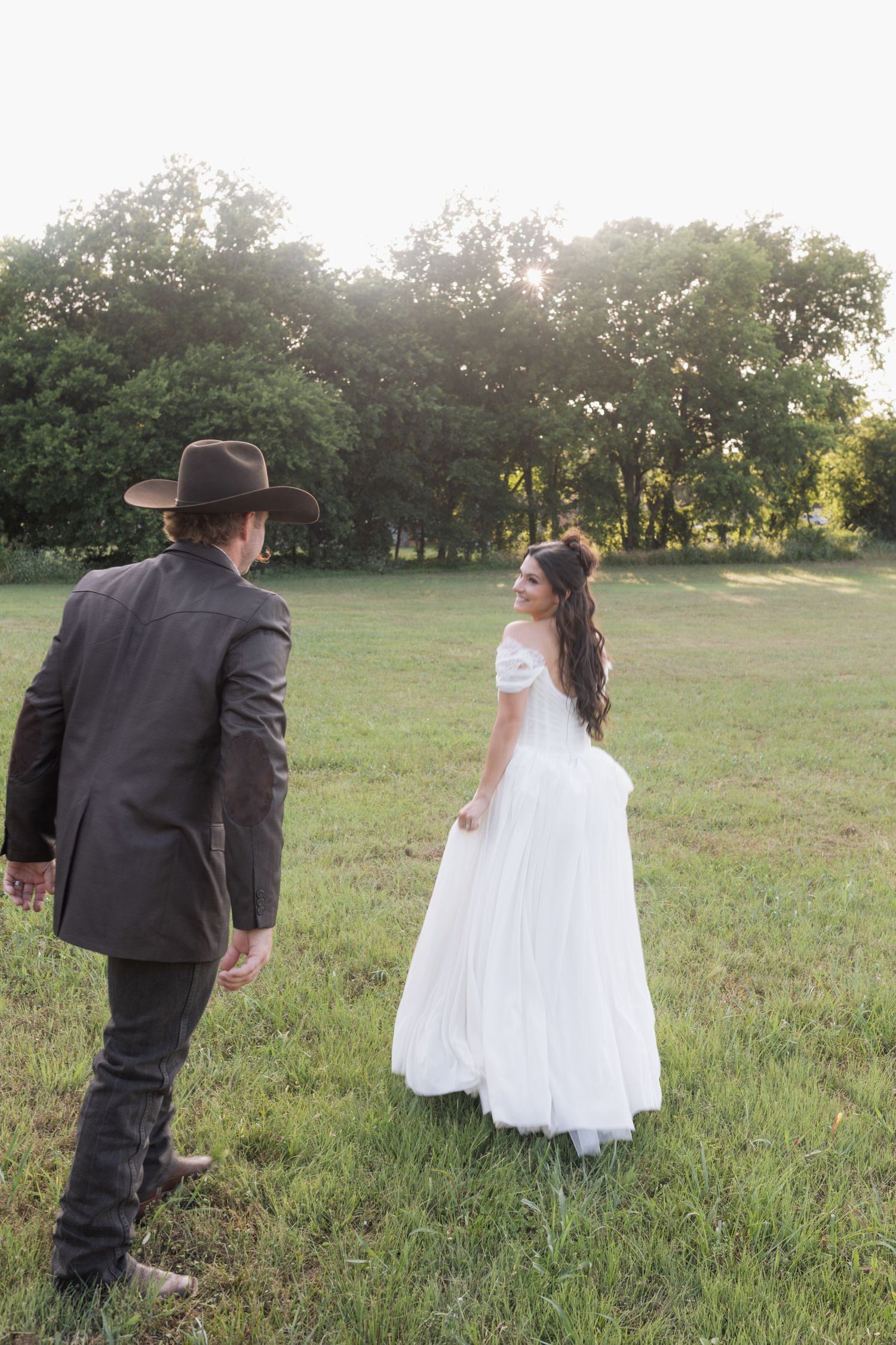 Bride in white gown looks back at groom wearing cowboy hat while walking through green meadow.
