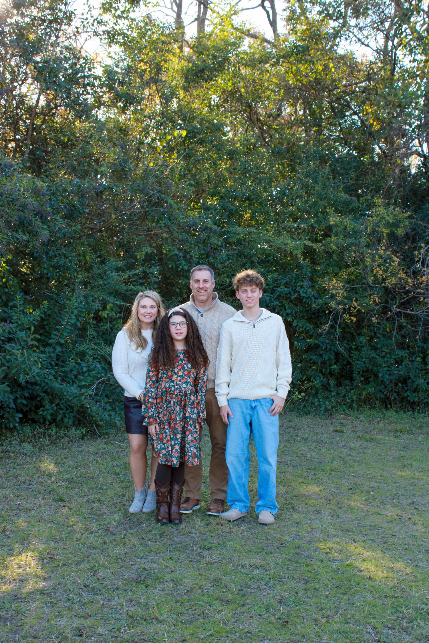 A group stands together outdoors in a grassy area with trees and bushes in the background during late afternoon light.