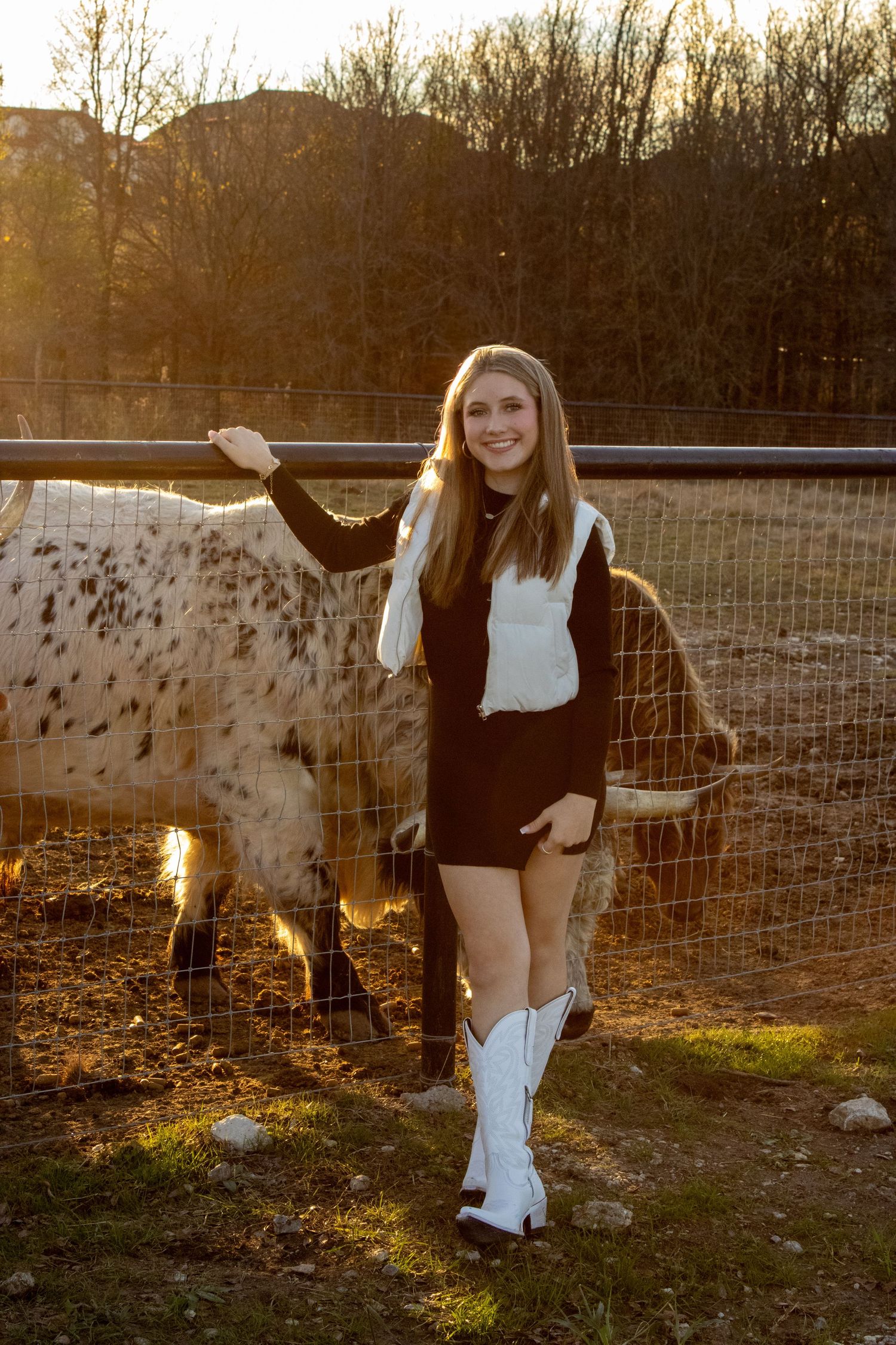 A spotted horse and person stand together in a fenced pasture during golden hour sunset lighting.