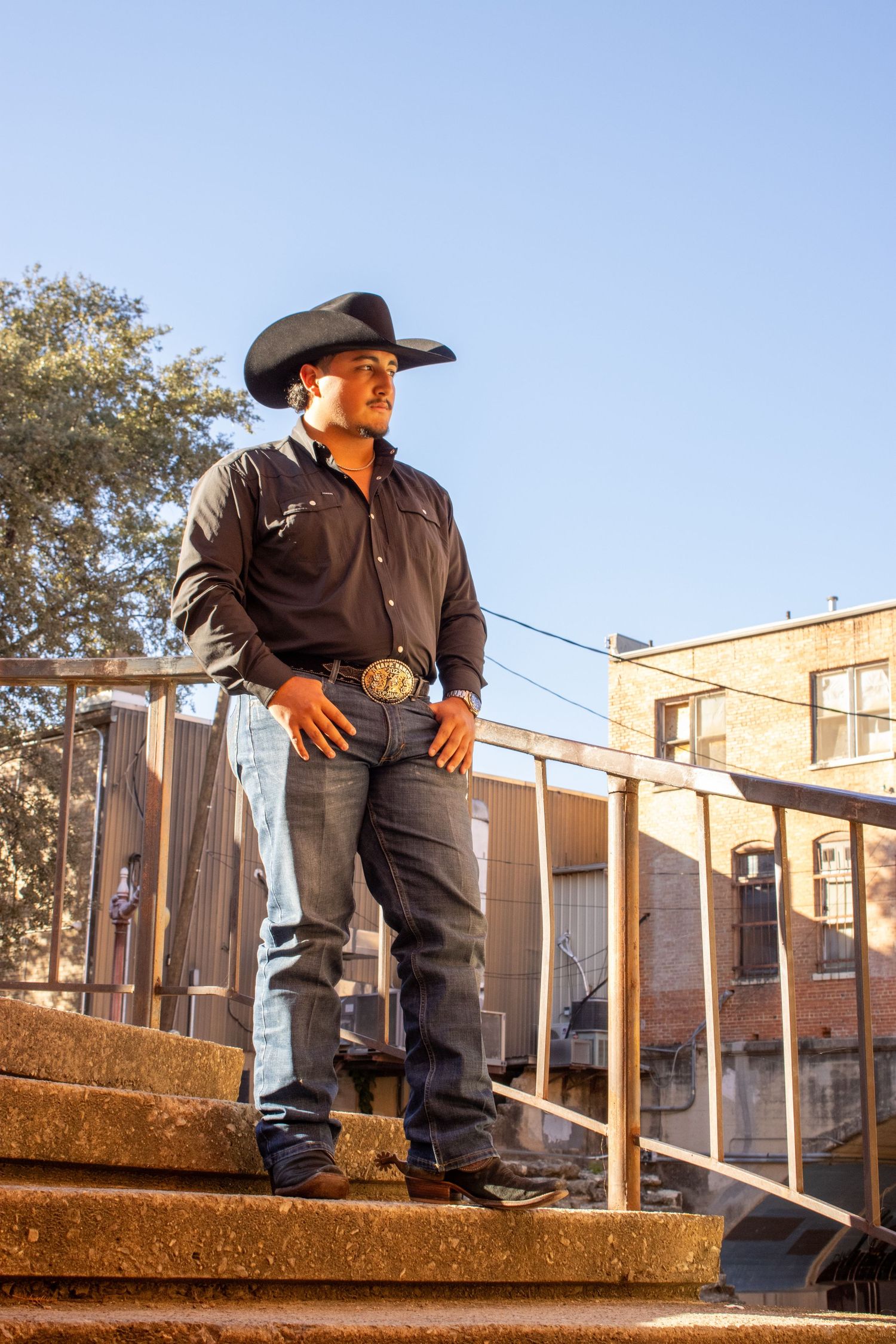 A cowboy in western attire stands confidently on concrete steps against an urban backdrop.