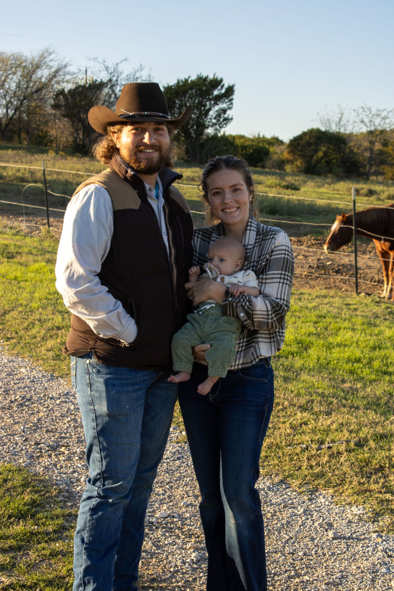 A couple stands together outdoors with a baby in a rural setting during golden hour.