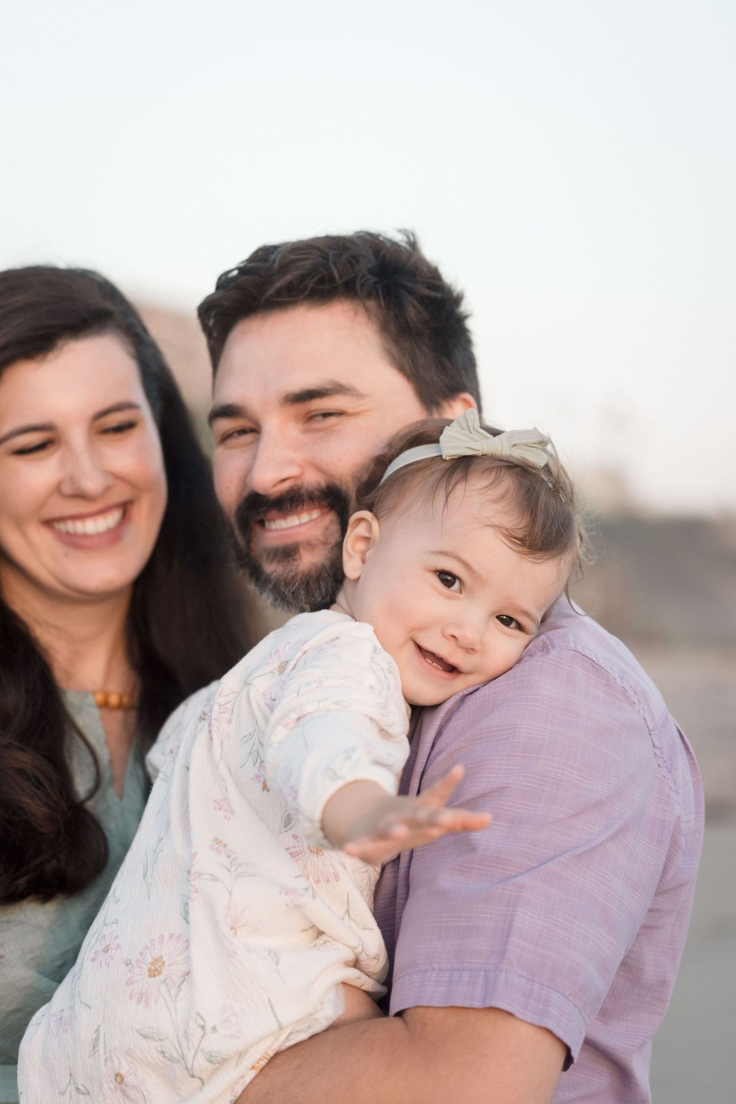 Joyful family closeup with bearded father holding smiling baby girl in white dress on beach.