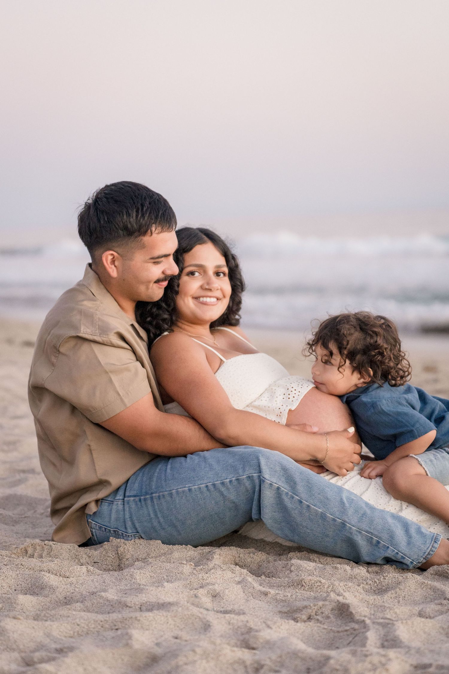 A loving family sits together on sandy beach, father embracing pregnant mother while toddler rests against her baby bump.