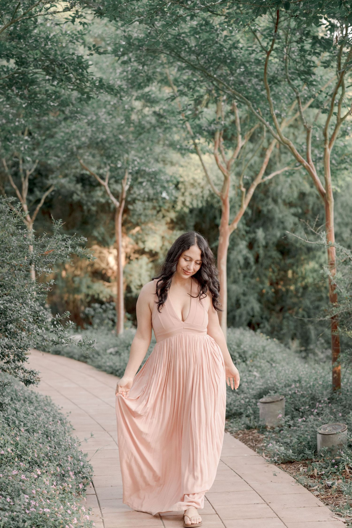 A woman in a flowing blush pink maxi dress walks along a tree-lined garden path.