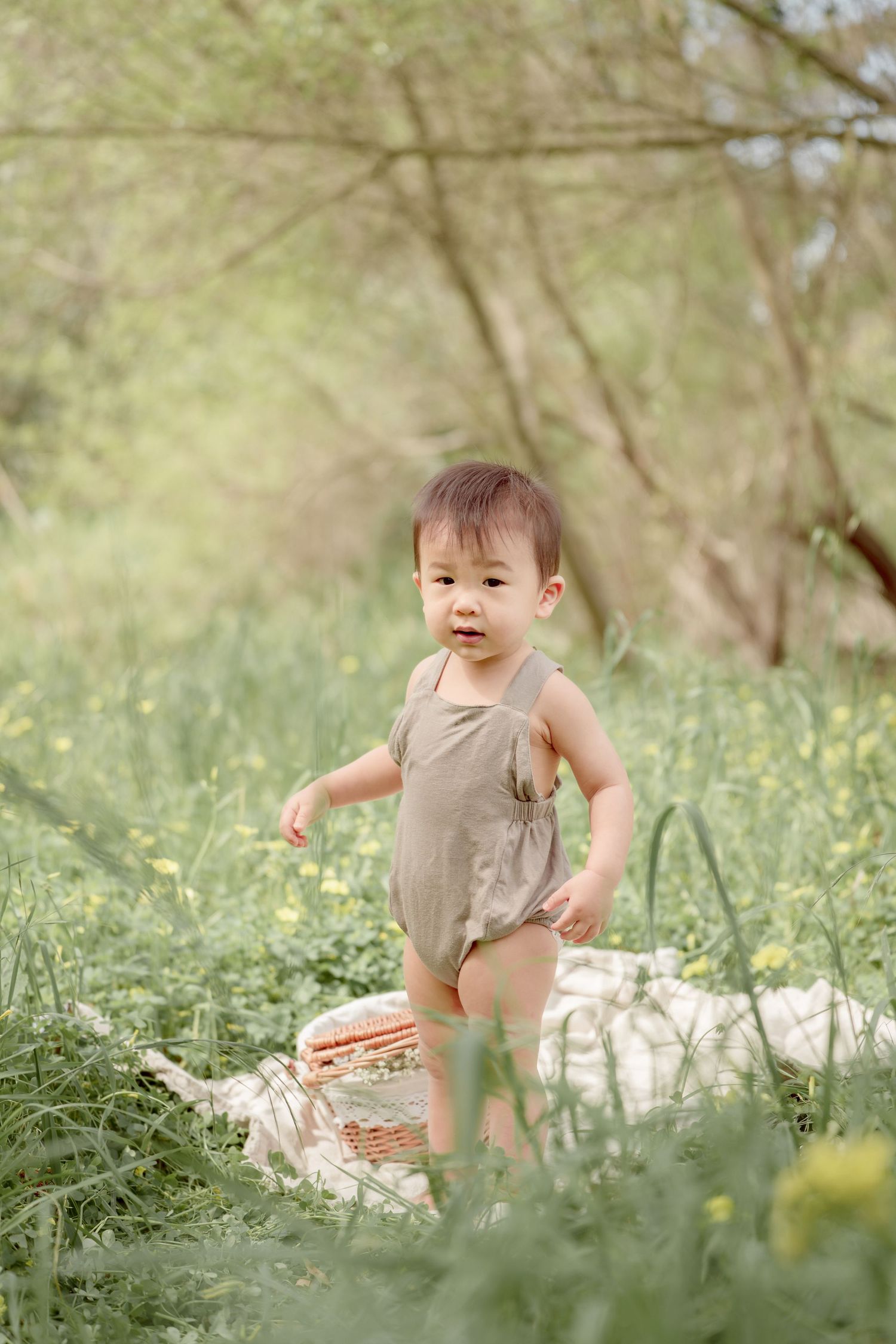 An Asian toddler in a green romper stands on a white blanket surrounded by tall grass and yellow wildflowers outdoors.
