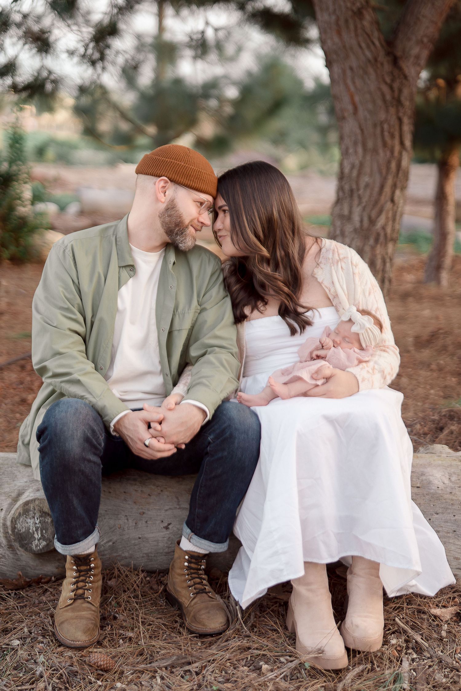Couple sitting on a log sharing a tender moment, man in green jacket and brown beanie, woman in white dress holding newborn.
