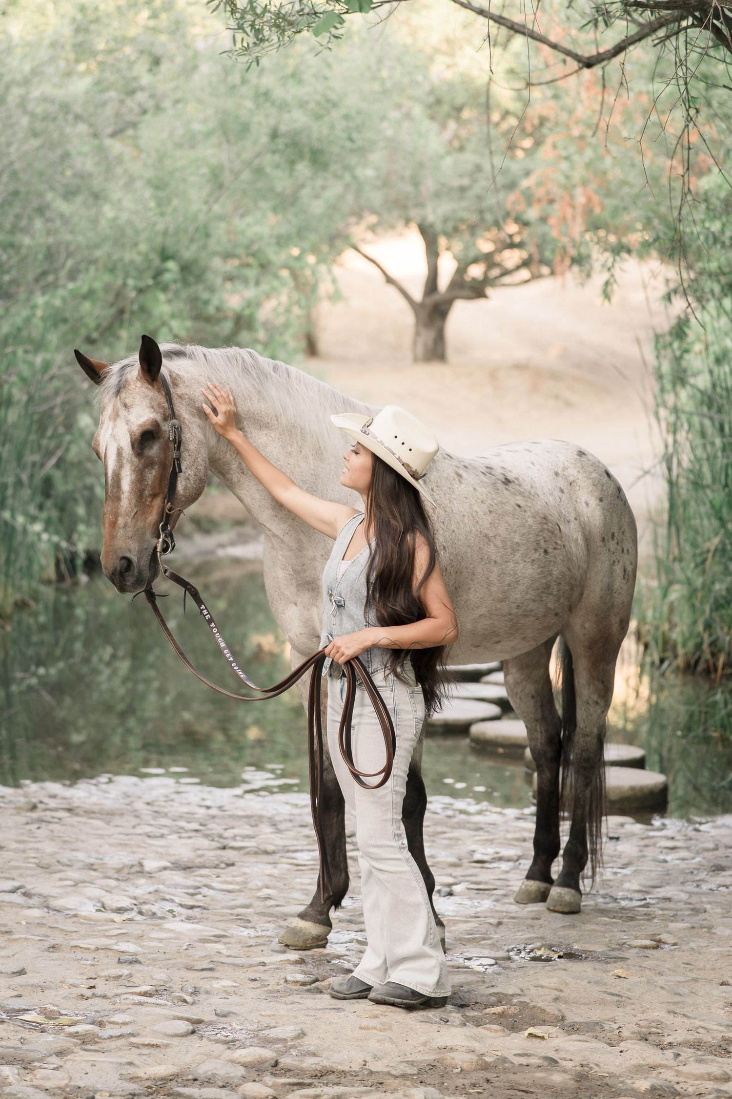 Woman in cowboy hat and linen outfit gently stroking a gray horse near a serene tree-lined path.