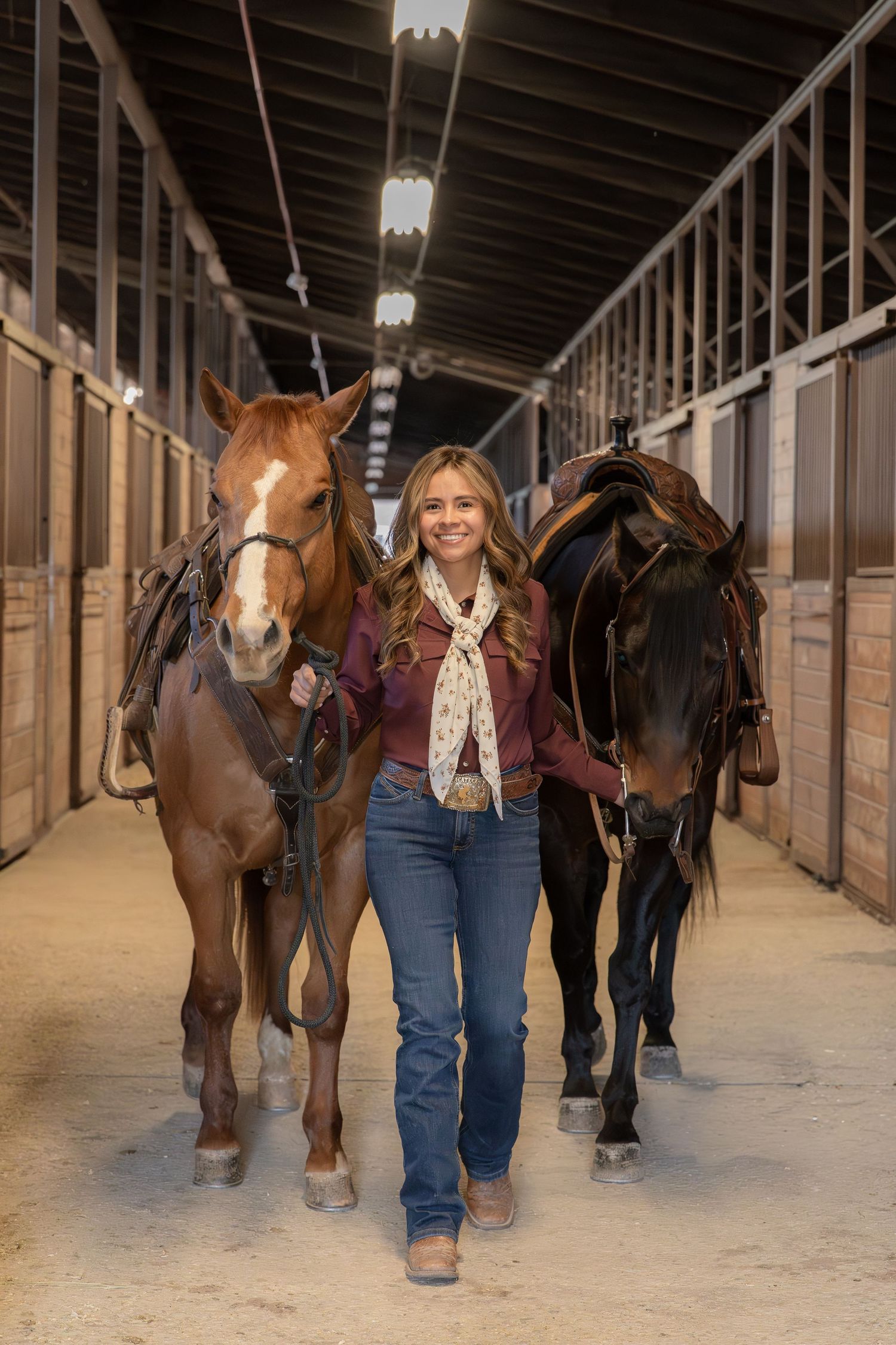 Young woman walking two saddled horses through a stable aisle, smiling confidently in jeans and a maroon top.