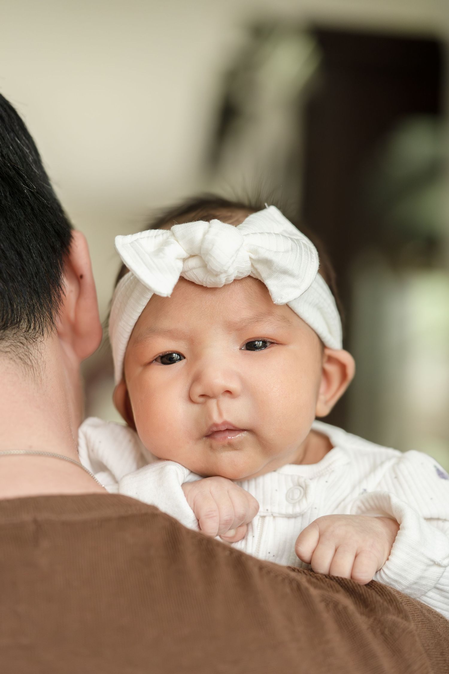 A newborn baby girl wearing a white bow headband rests on a parent's shoulder during a newborn photo session.
