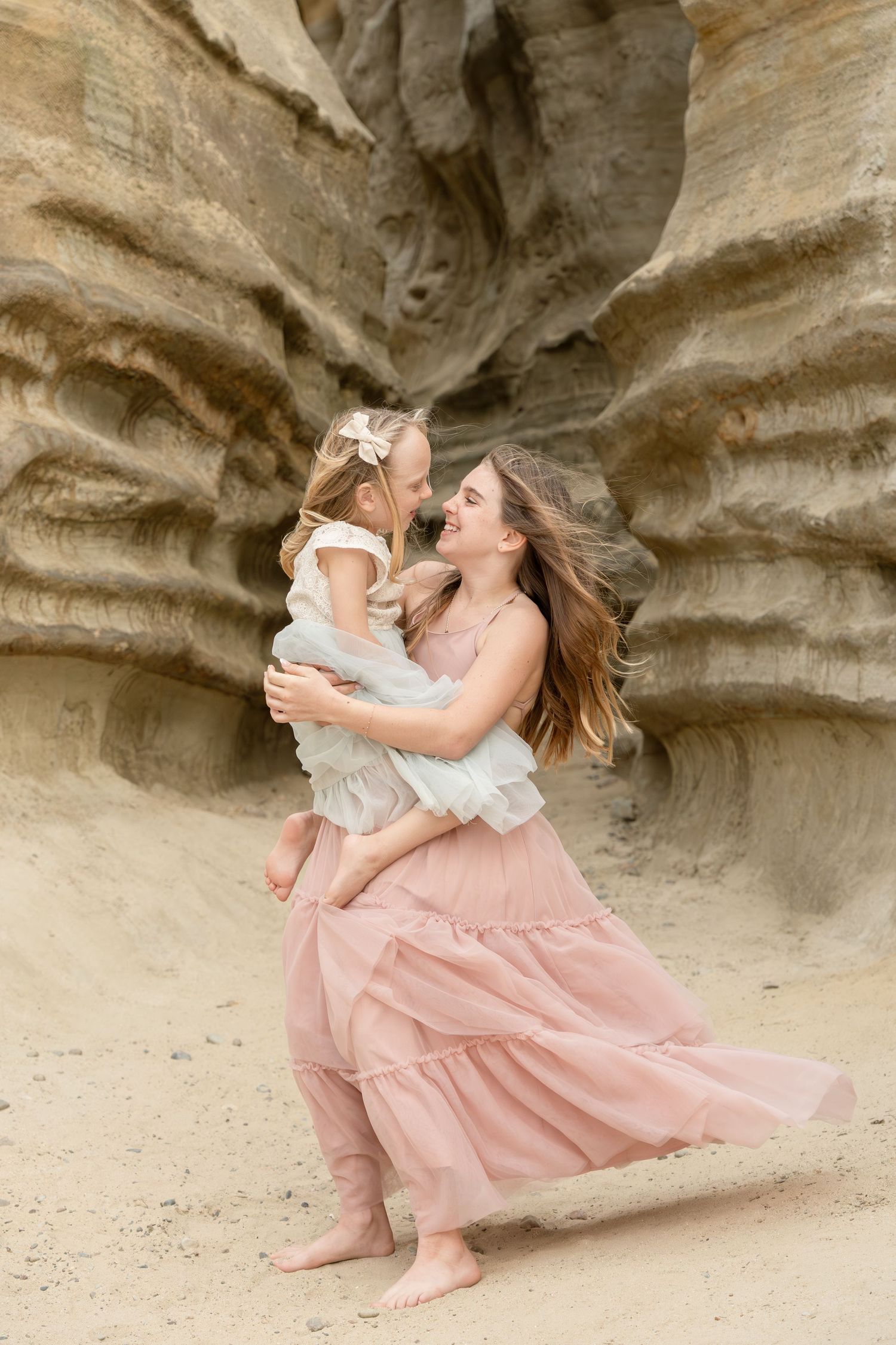 Mother in pink dress twirls barefoot daughter in white dress before sandy rock formations.