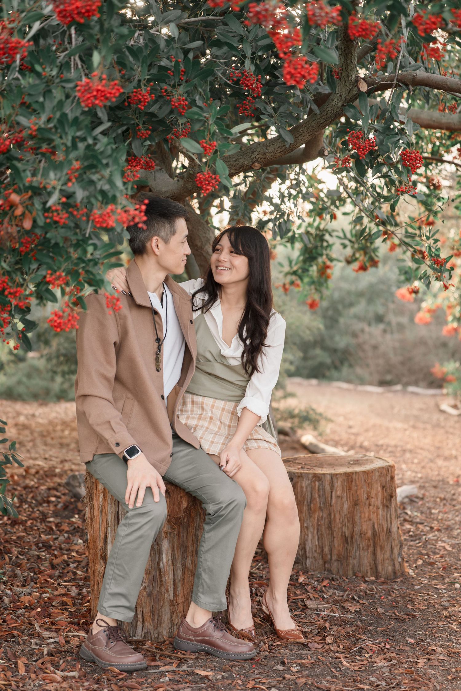 A couple sits romantically on a tree stump beneath a vibrant red berry tree in an autumn park setting.