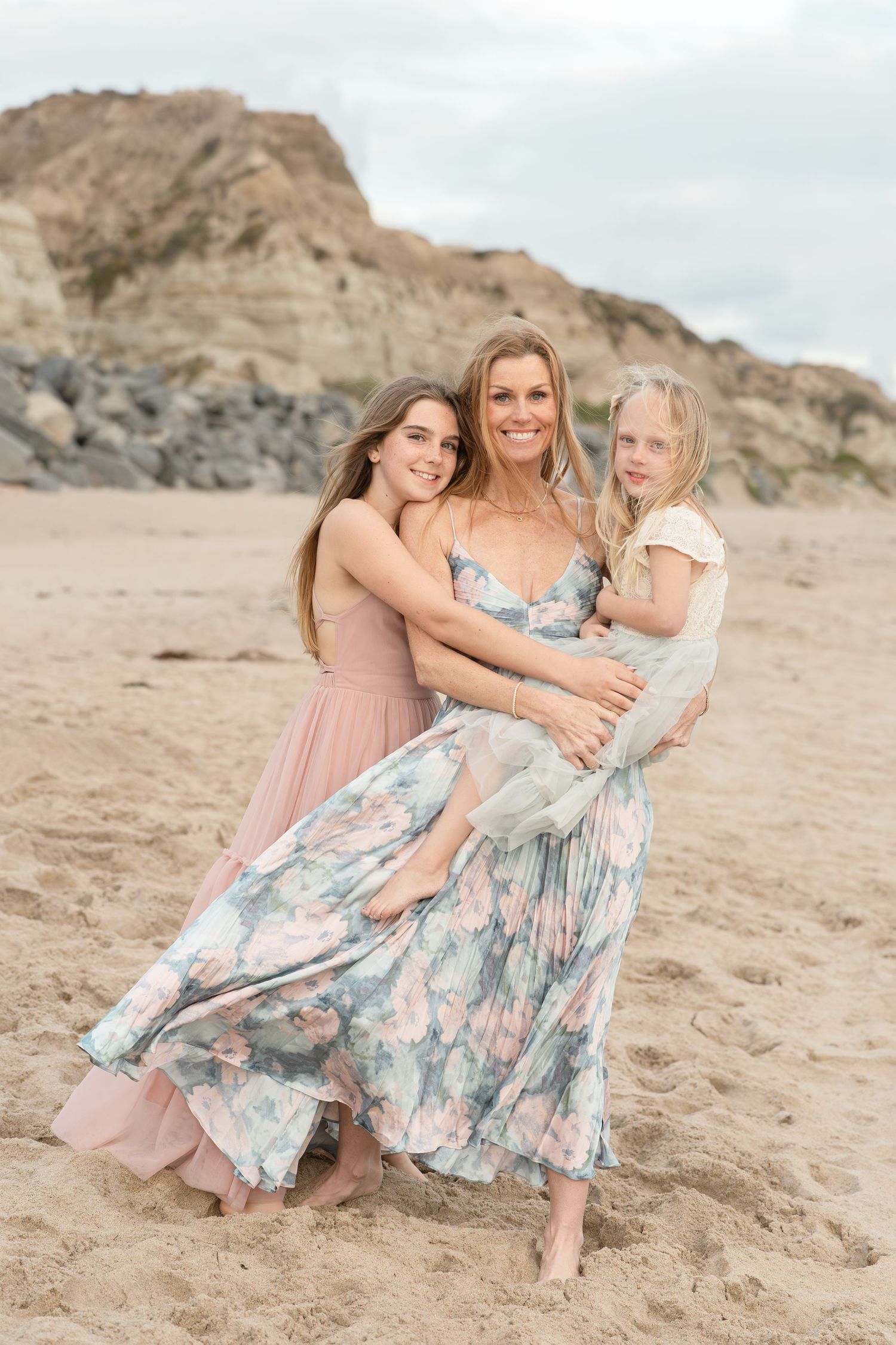 Mother in floral dress embracing two daughters on sandy beach with rocky cliffs.