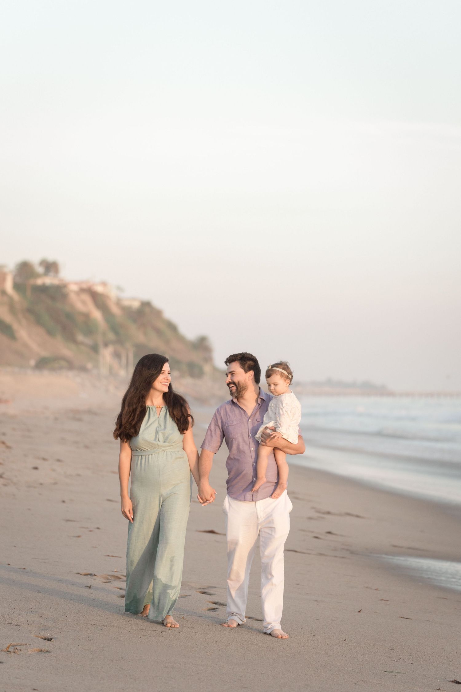 Happy family walking on beach at sunset, bearded dad carrying toddler while holding moms hand.