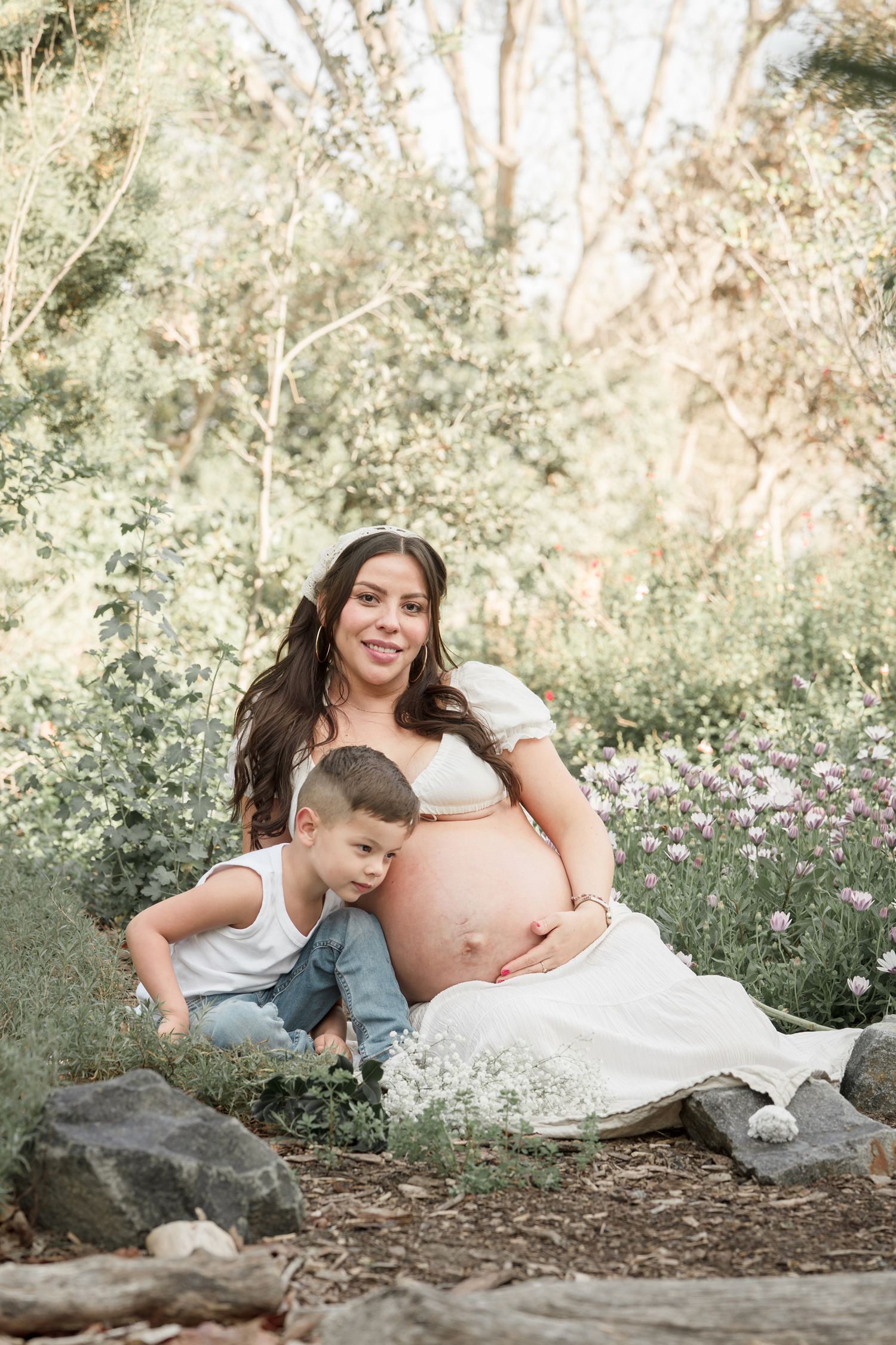 Pregnant woman in white dress sits outdoors with young boy leaning on her belly, surrounded by wildflowers and trees.