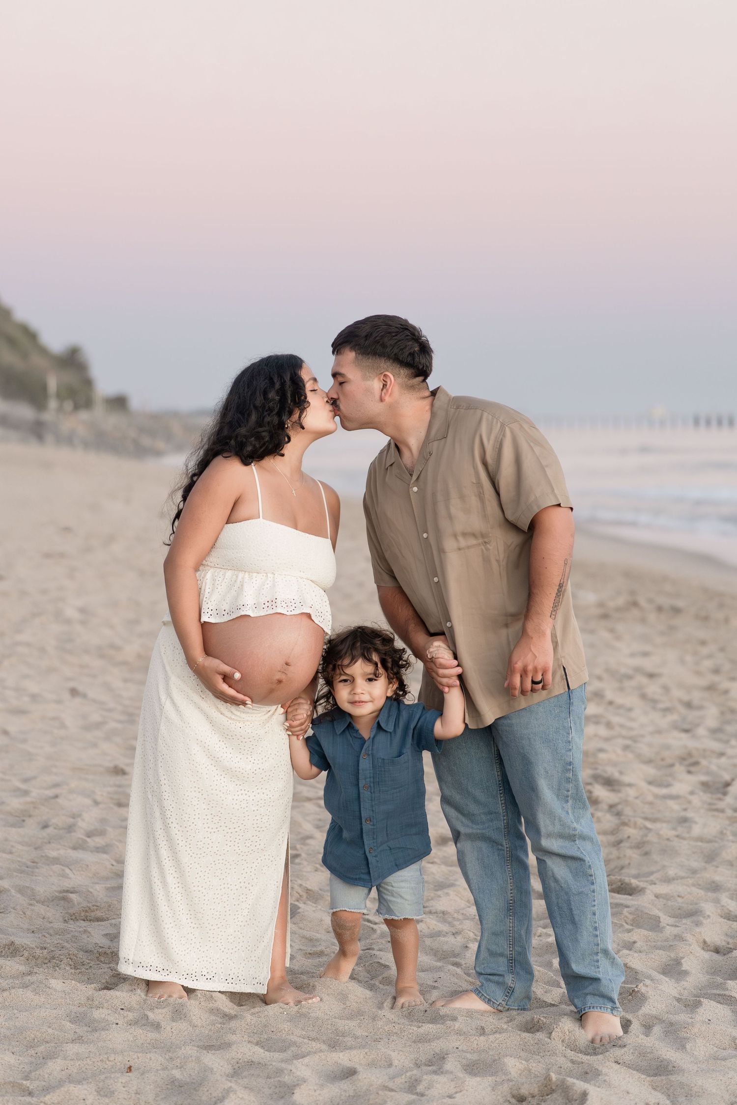 A pregnant couple kisses on the beach at sunset while their toddler son holds their hands.
