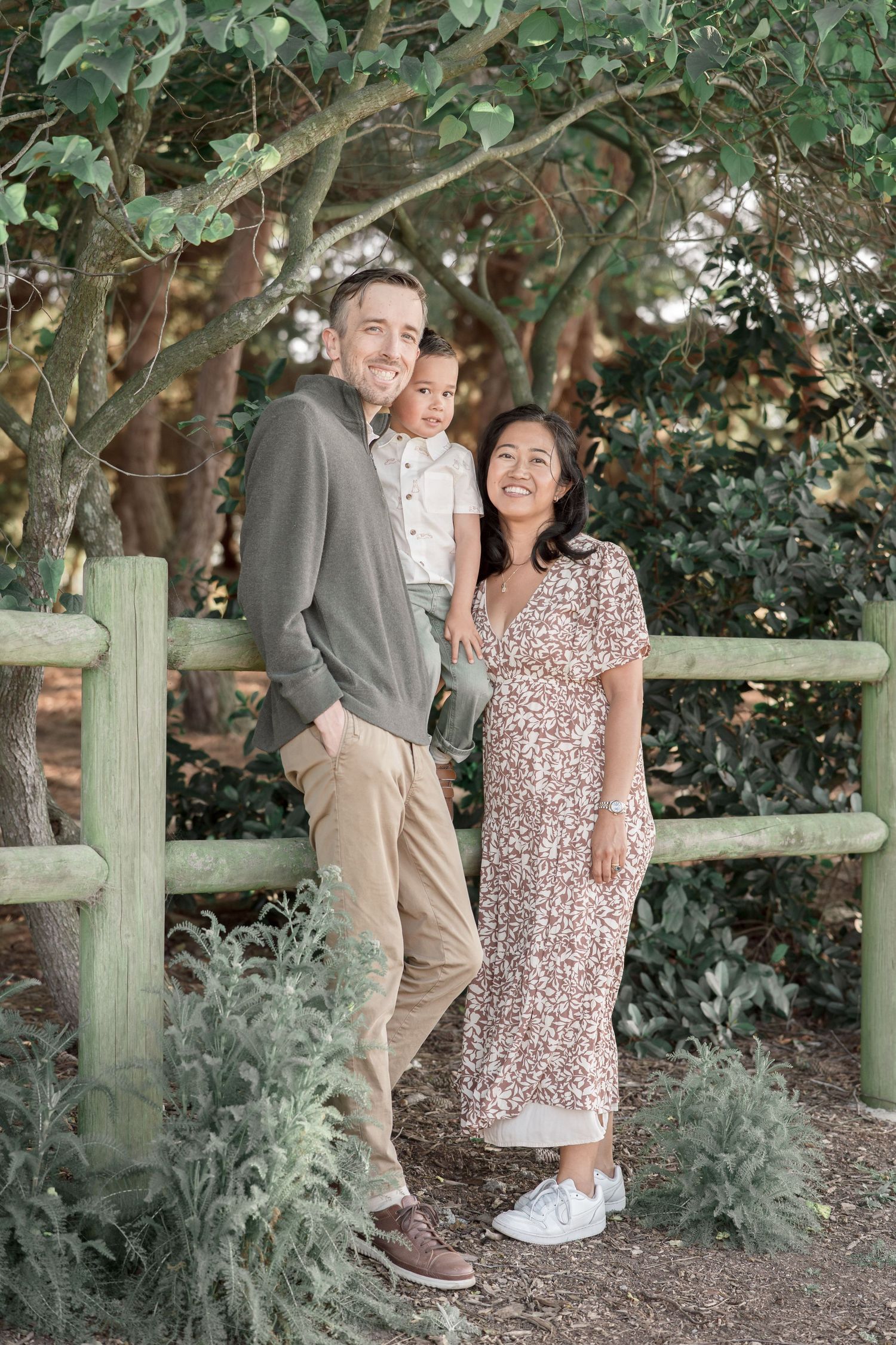 A smiling family of three poses outdoors by a wooden fence surrounded by lush green trees and plants.