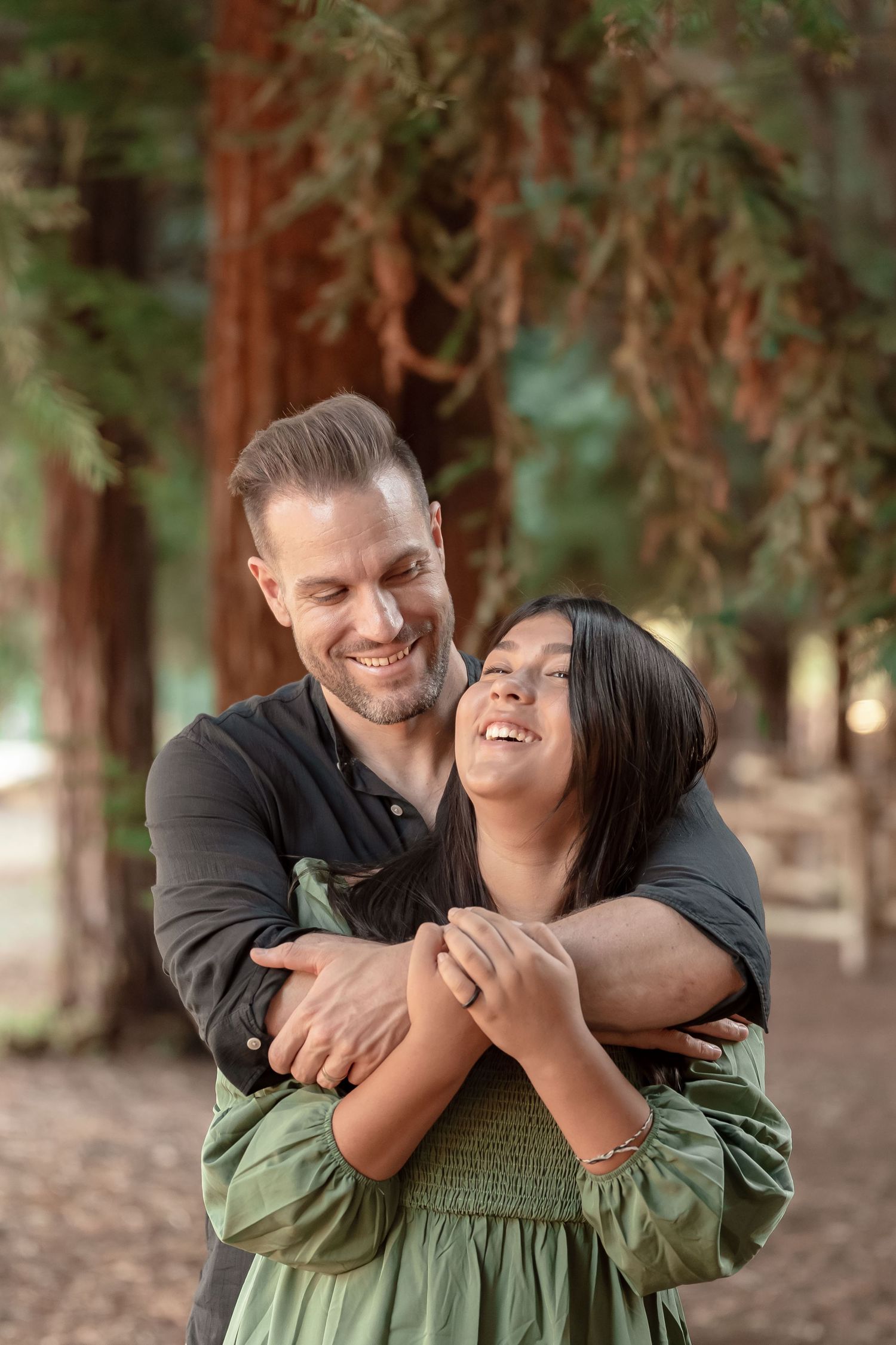 Smiling couple embracing warmly among tall redwood trees, man hugging woman from behind outdoors.