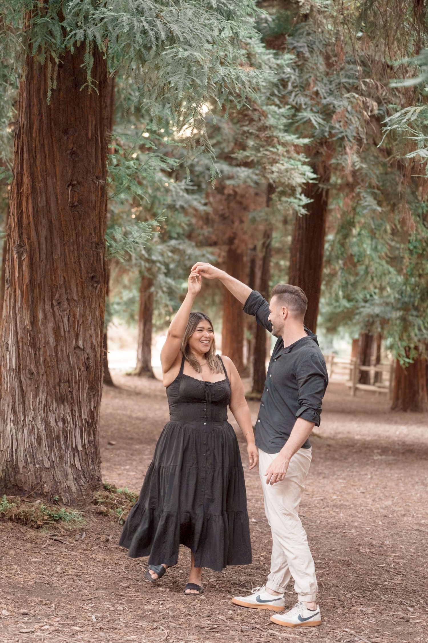 Couple dancing joyfully among tall redwood trees, woman in black dress being twirled by partner.
