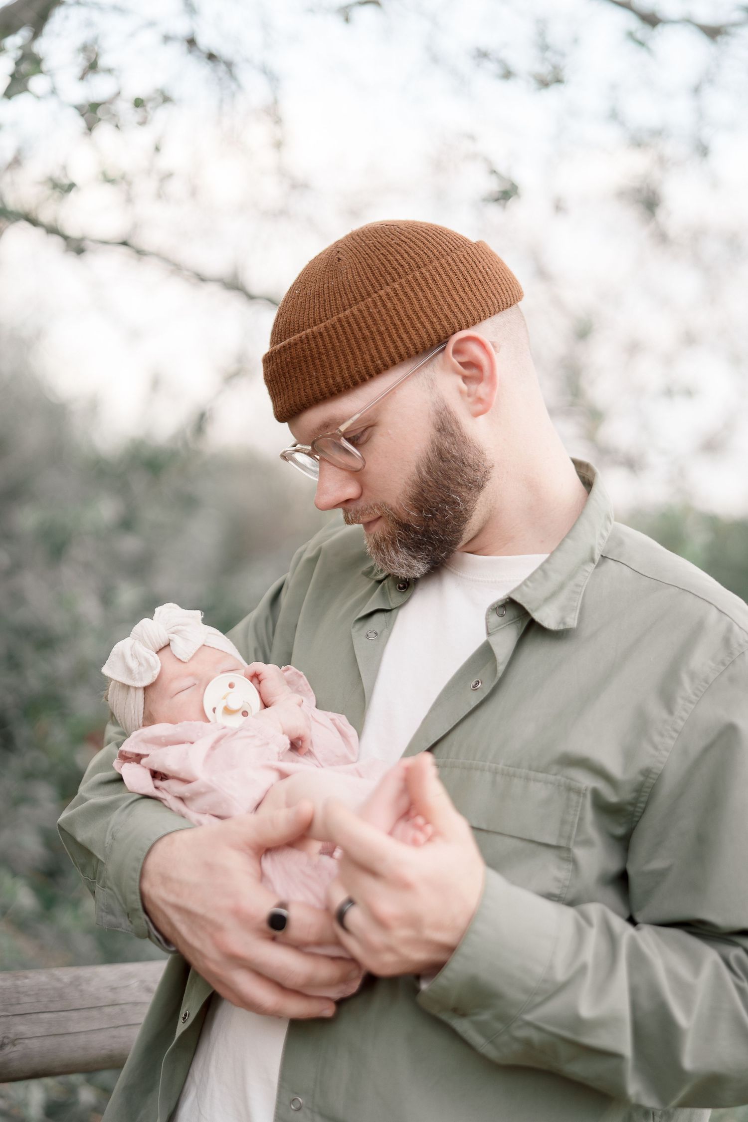 Bearded father in green jacket and brown beanie tenderly holding newborn baby outdoors among blooming trees.